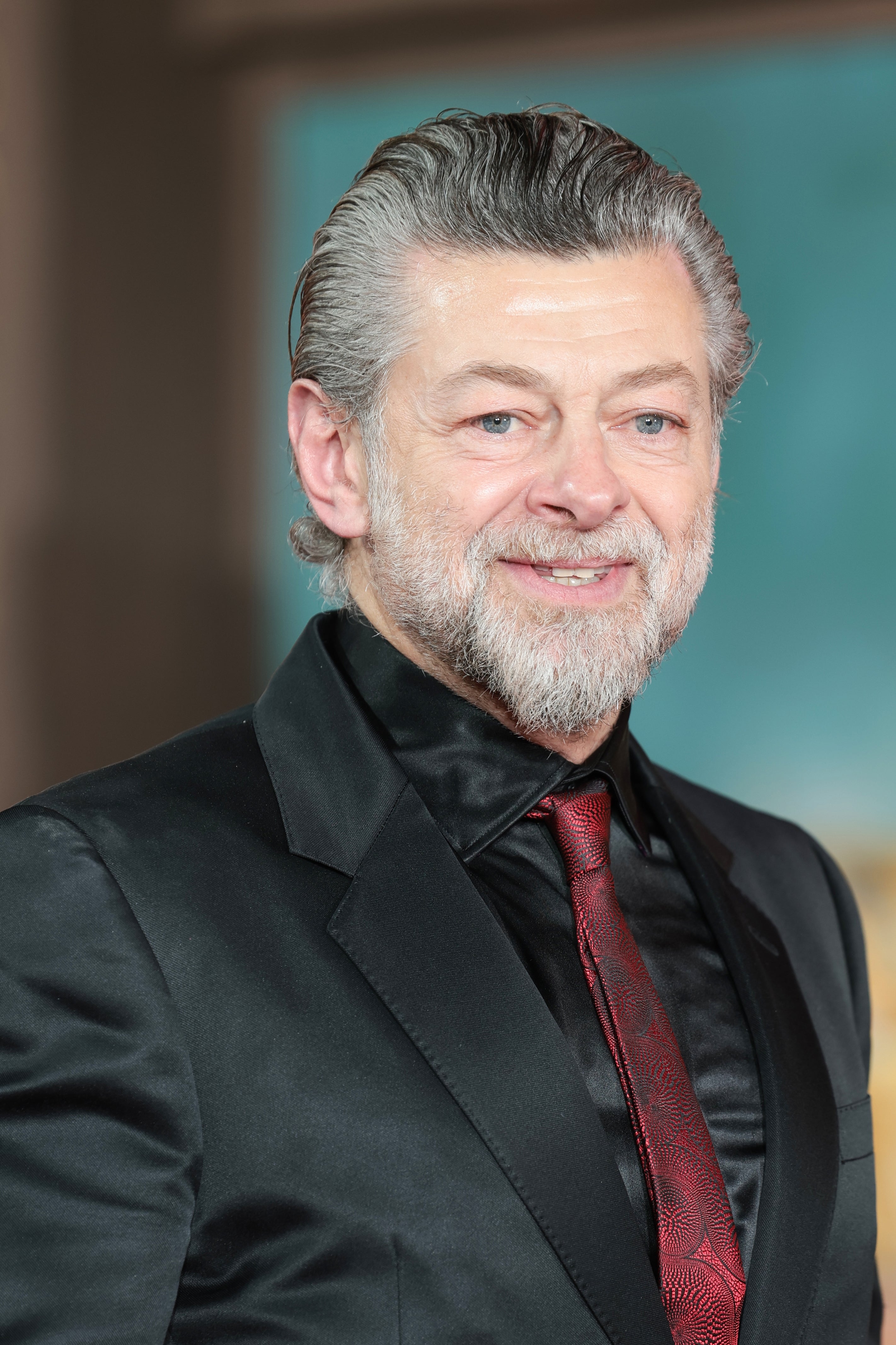 A person in a sleek black suit with a patterned red tie smiles at a formal event