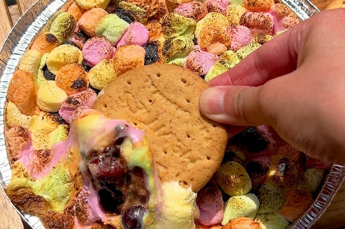 手がビスケットを持ち、焼きマシュマロと溶けたチョコの入ったスイーツにディップしている。