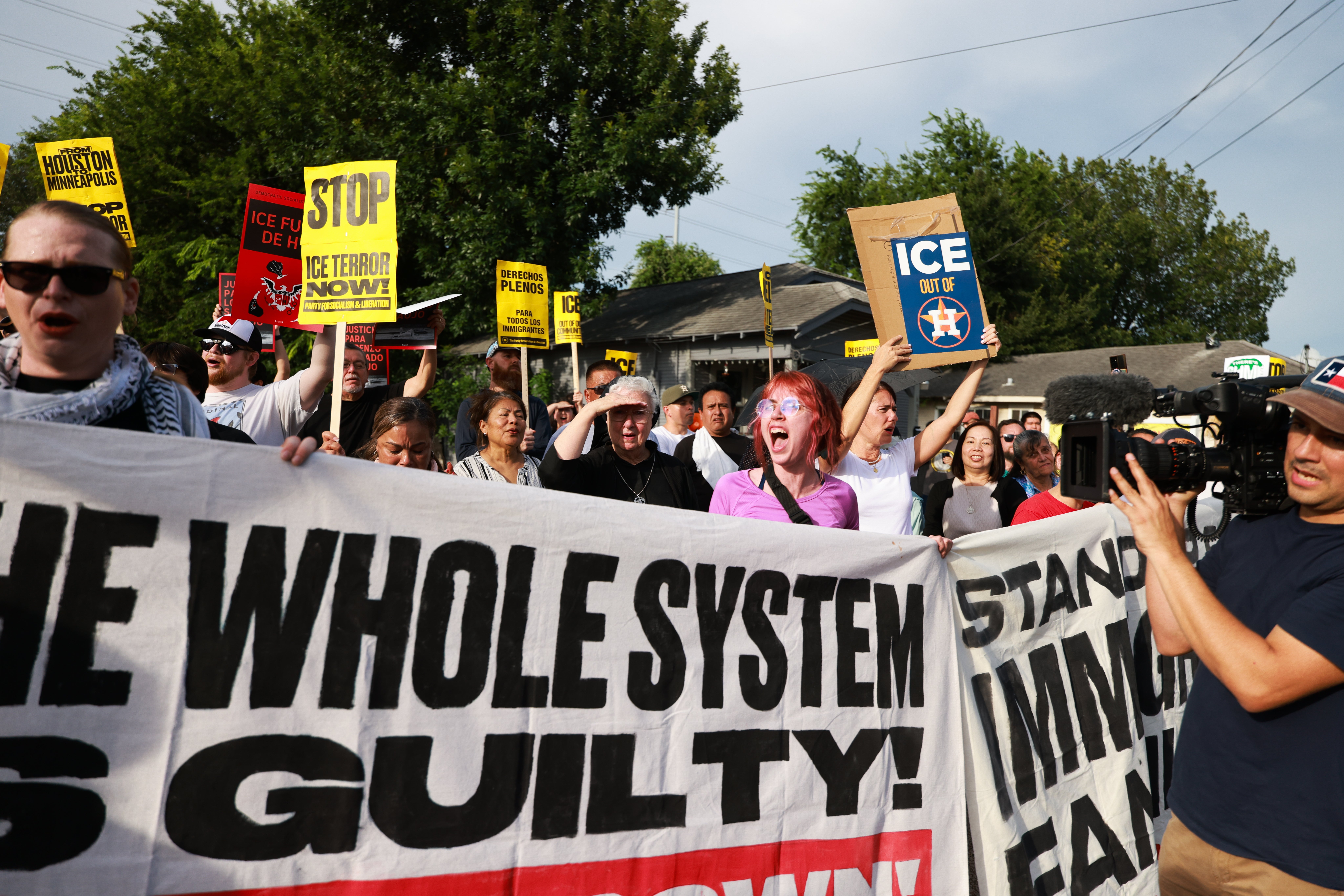 Protesters march with signs against ICE and the migration system, displaying messages similar "The Whole System is Guilty" and "Stop ICE Terror Now."