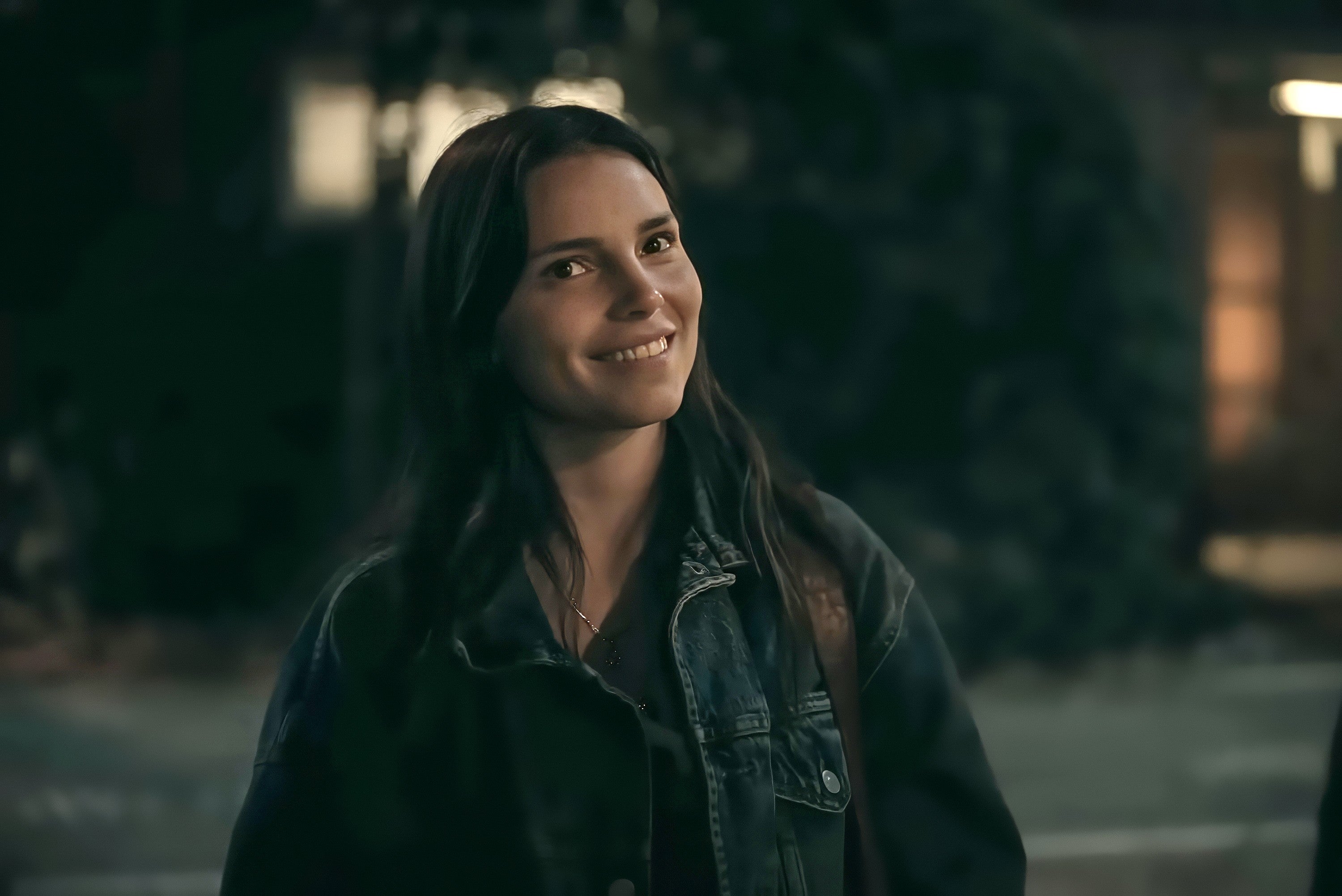 A person smiling outdoors at night, wearing a denim jacket and a backpack