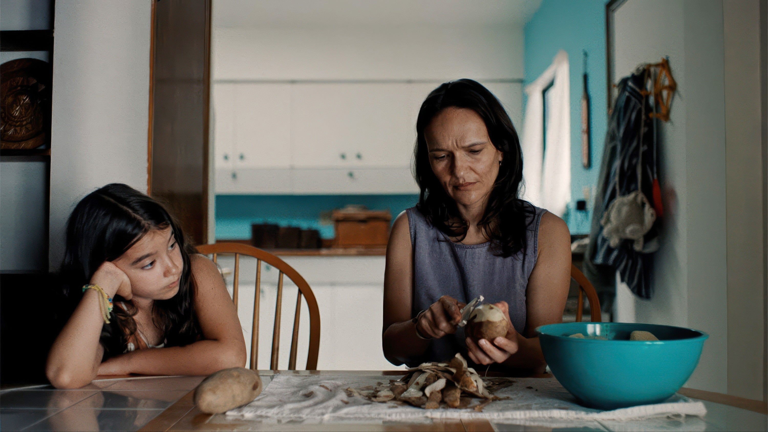 Woman peeling potatoes at a table; young girl next to her looks on thoughtfully. Kitchen setting in background