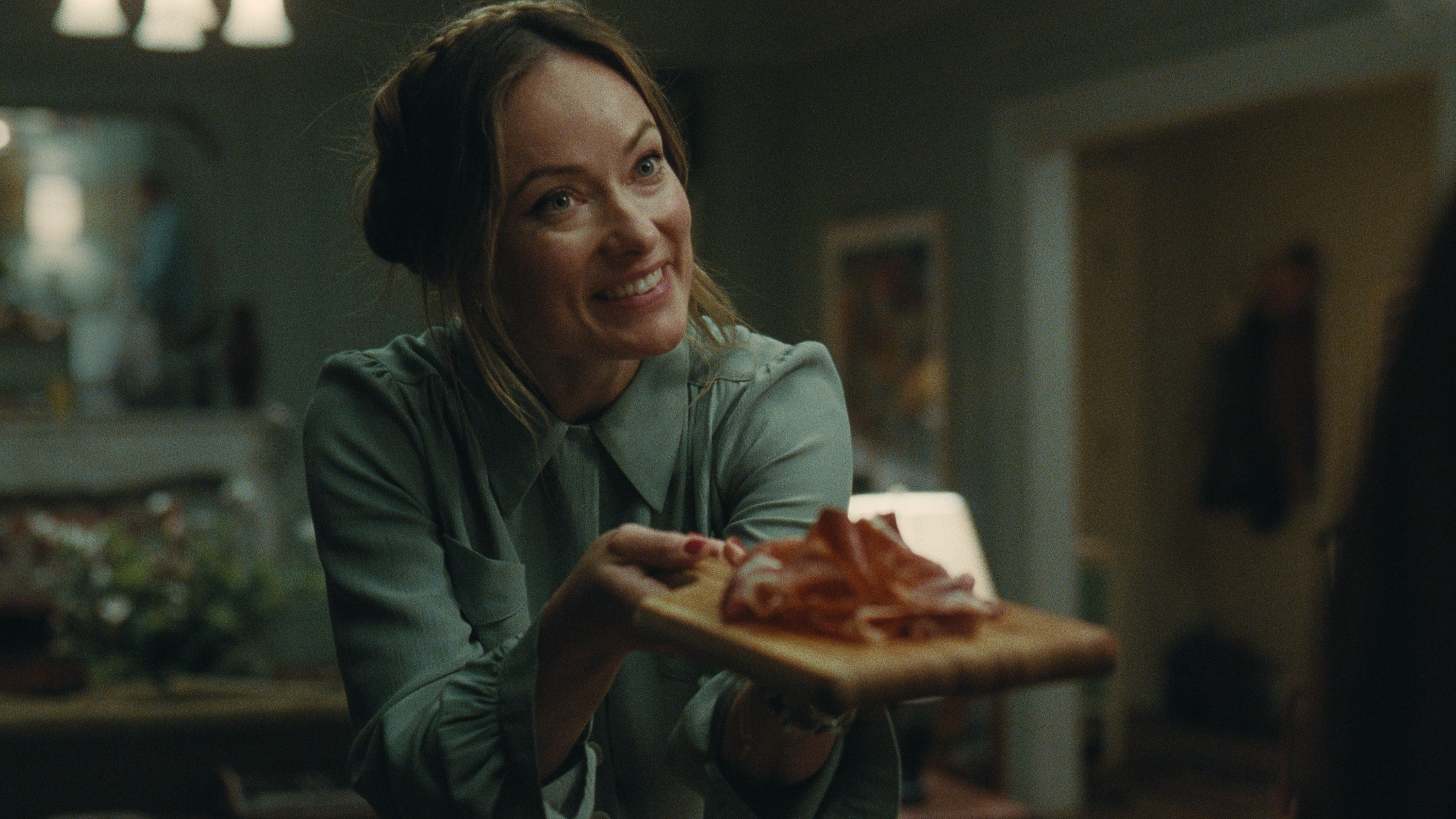 A smiling woman in a blouse offers a cutting board with neatly arranged slices of food, likely during a social gathering at home