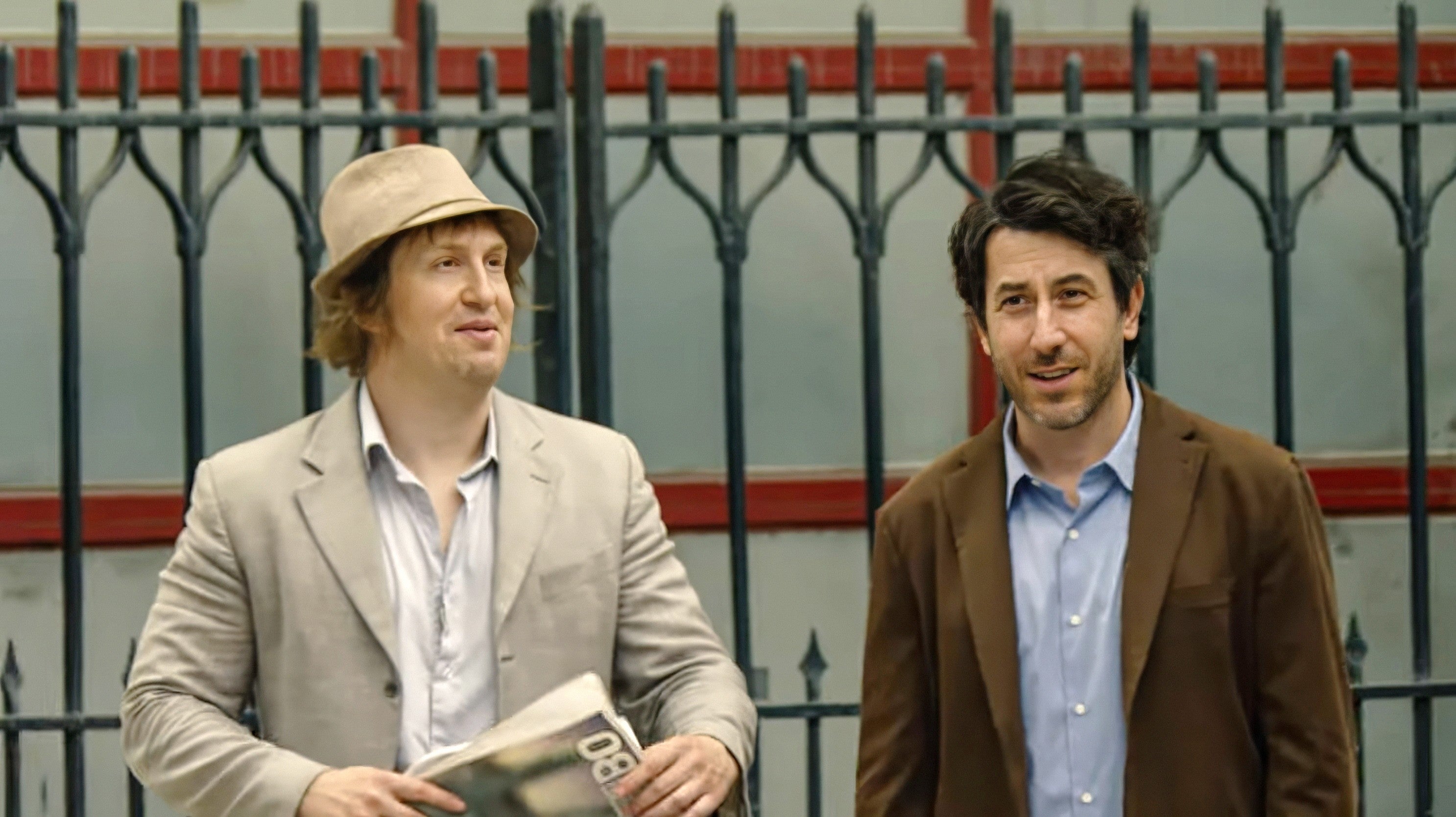 Two men in suits, one holding a newspaper, stand in front of a wrought iron fence