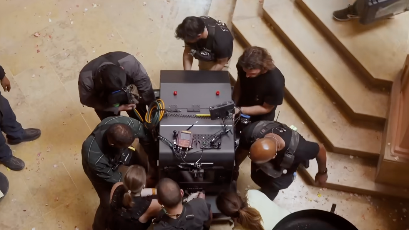 Group of people surrounding and working on a large electronic device indoors, near a staircase. Technical setup scene