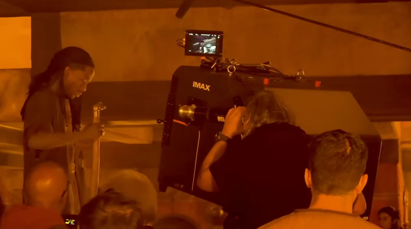 Film crew captured in action, operating an IMAX camera in a bustling scene