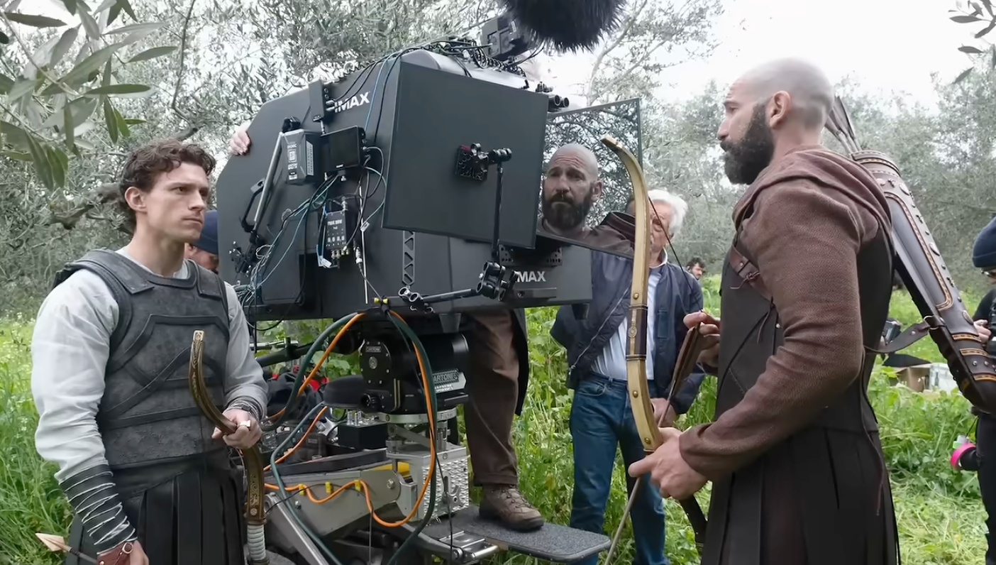 Actors on a film set in medieval-style costumes with bows, standing beside a large camera setup outdoors