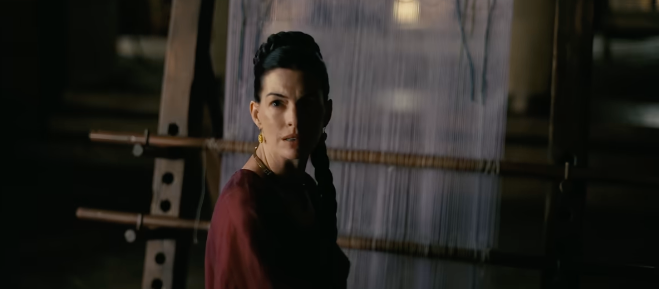 Woman with braided hair in a dimly lit room, looking towards the camera with a serious expression. Background is blurred