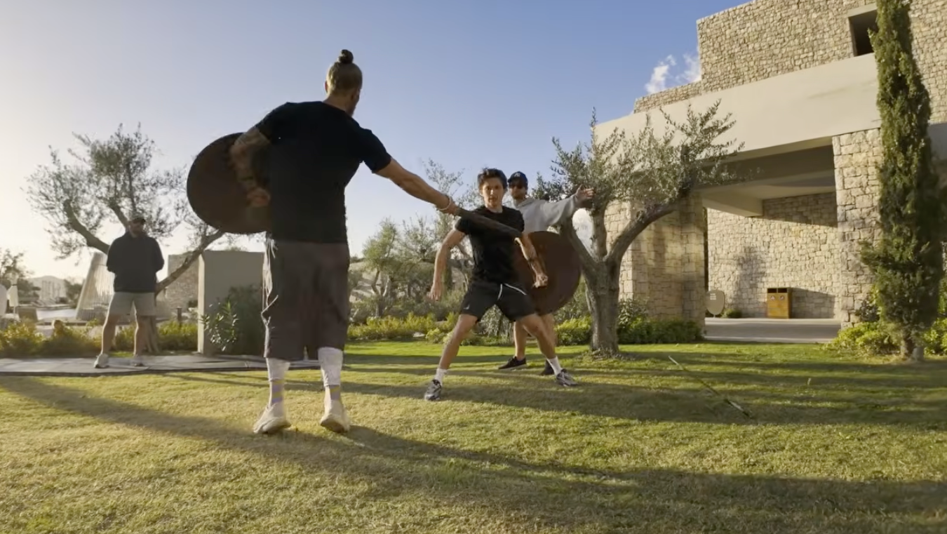 People practicing sword fighting outdoors in a yard, surrounded by trees and a large building