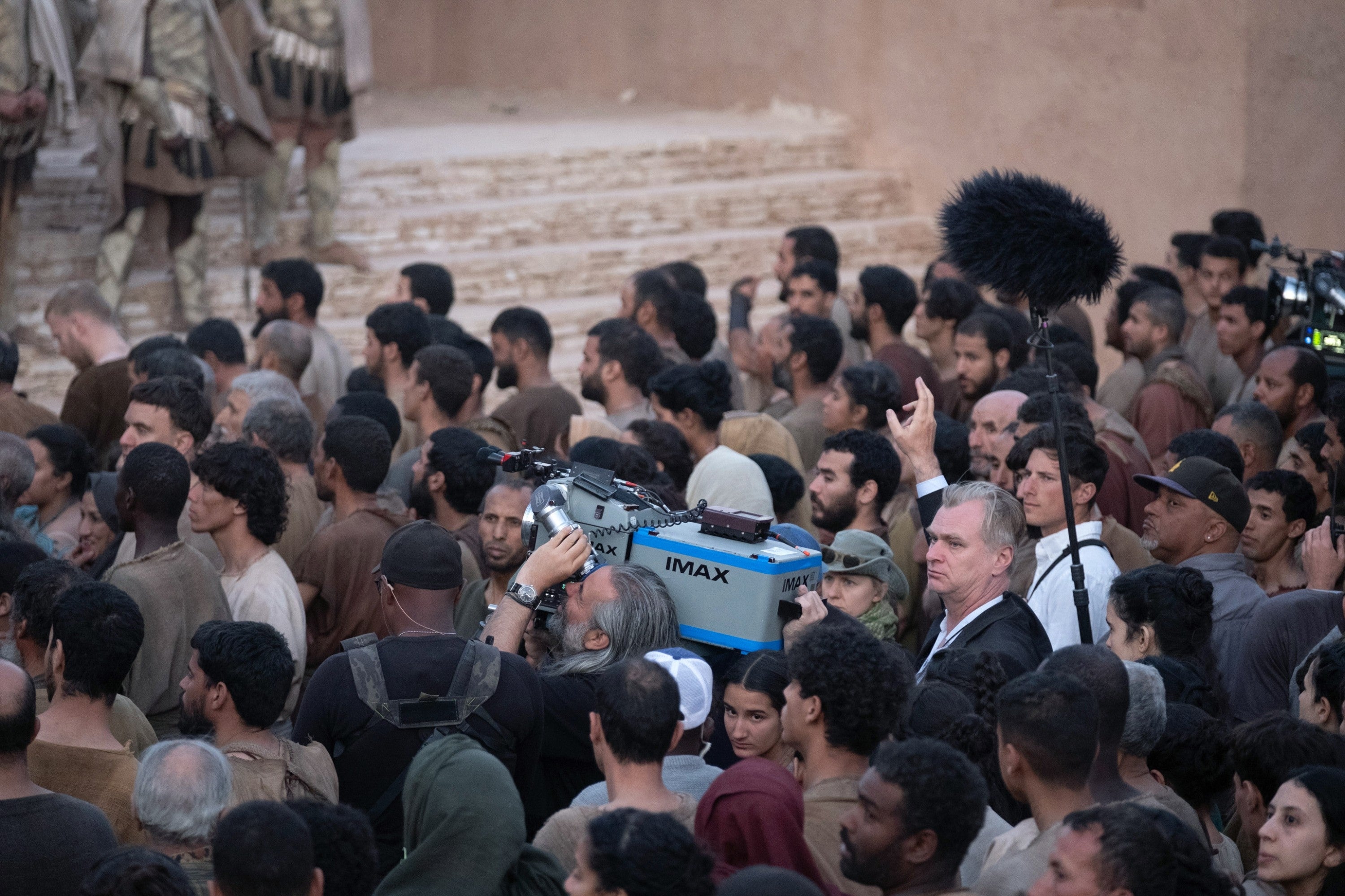 A film crew with an IMAX camera is capturing a scene in a crowd of people dressed in historical attire, some crew in modern gear