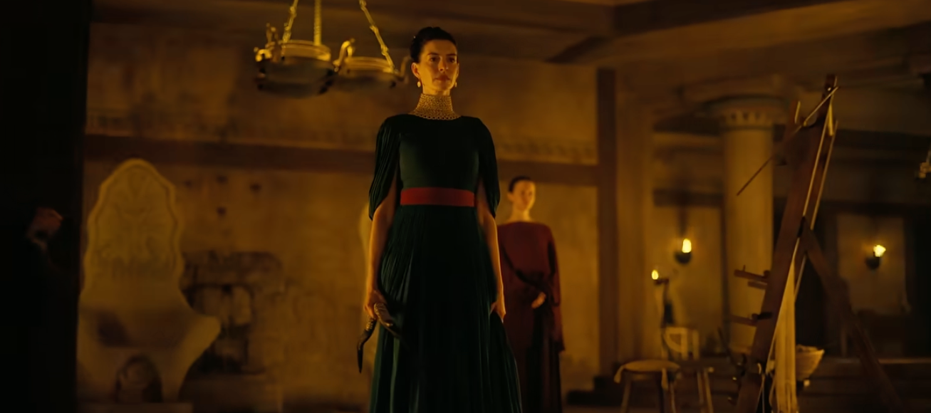 Two women in a dimly lit, elegant room; one wears a long dress with a red belt