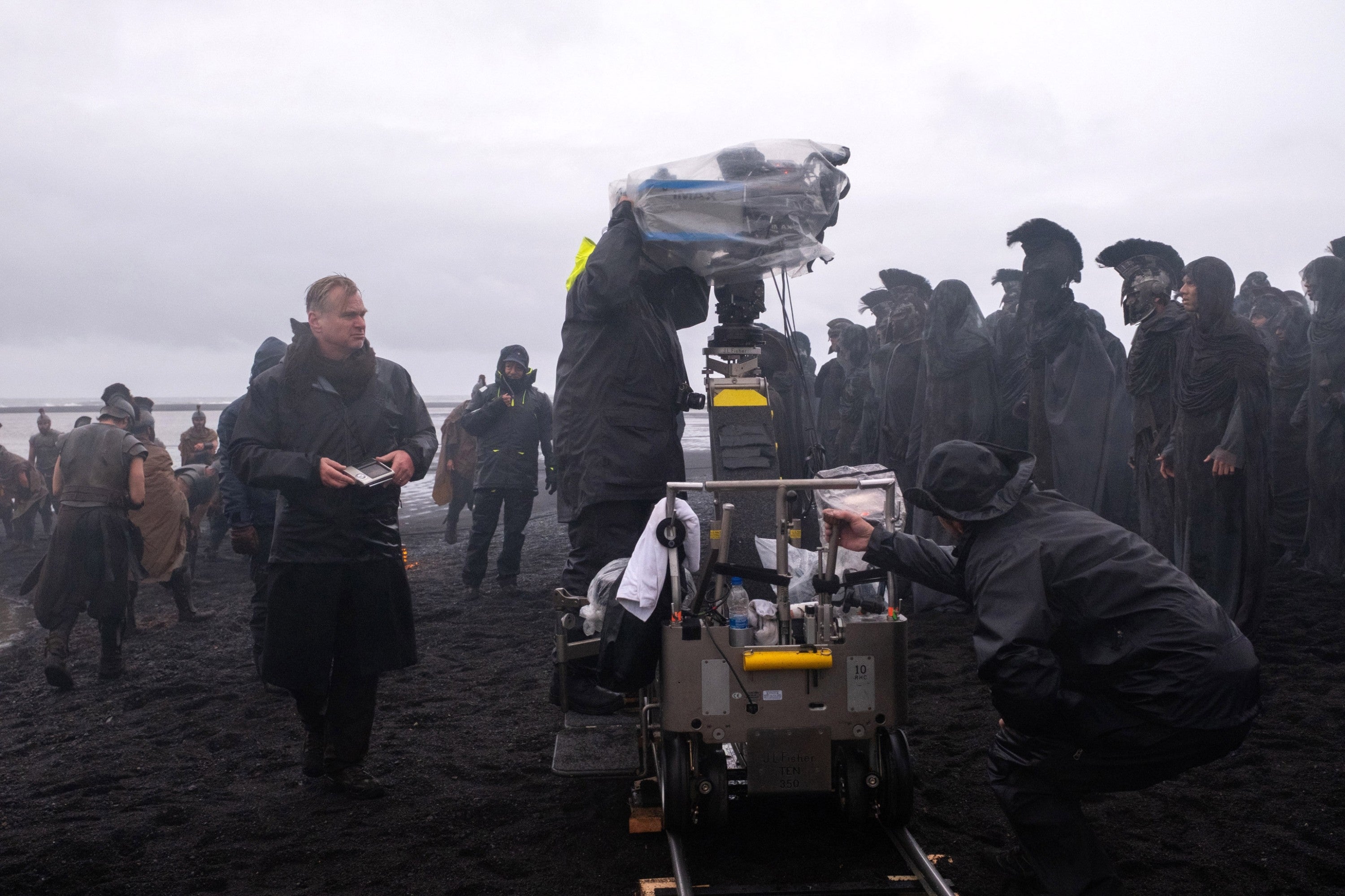 Film crew on a beach set; members in weatherproof gear operate equipment, surrounded by people in period costumes