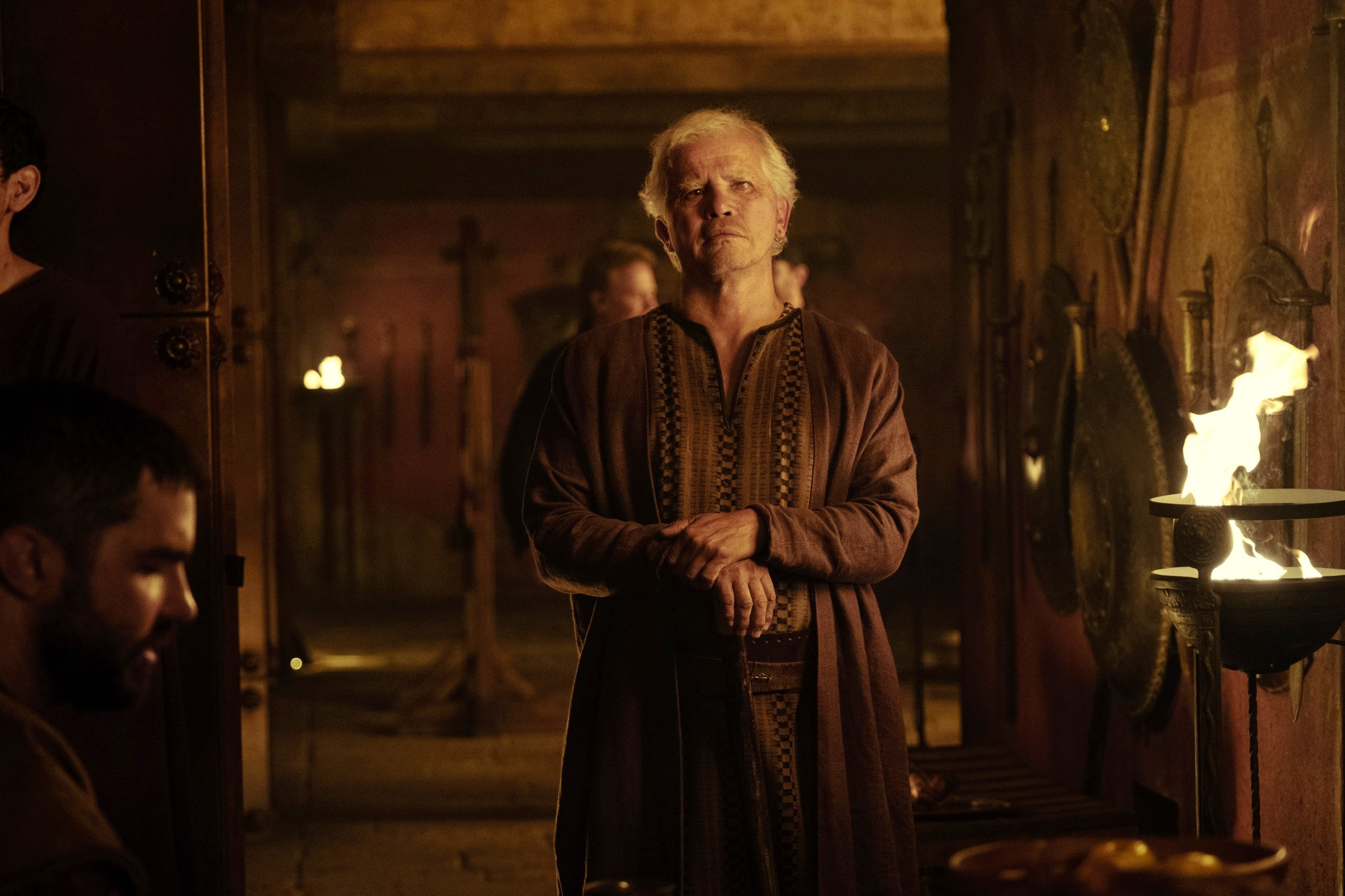 Elderly man in medieval-style robe stands in dimly lit room with torches