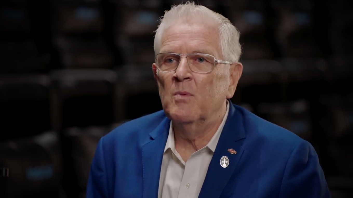 An older adult with short white hair and glasses is wearing a blue blazer with pins, speaking in a formal setting