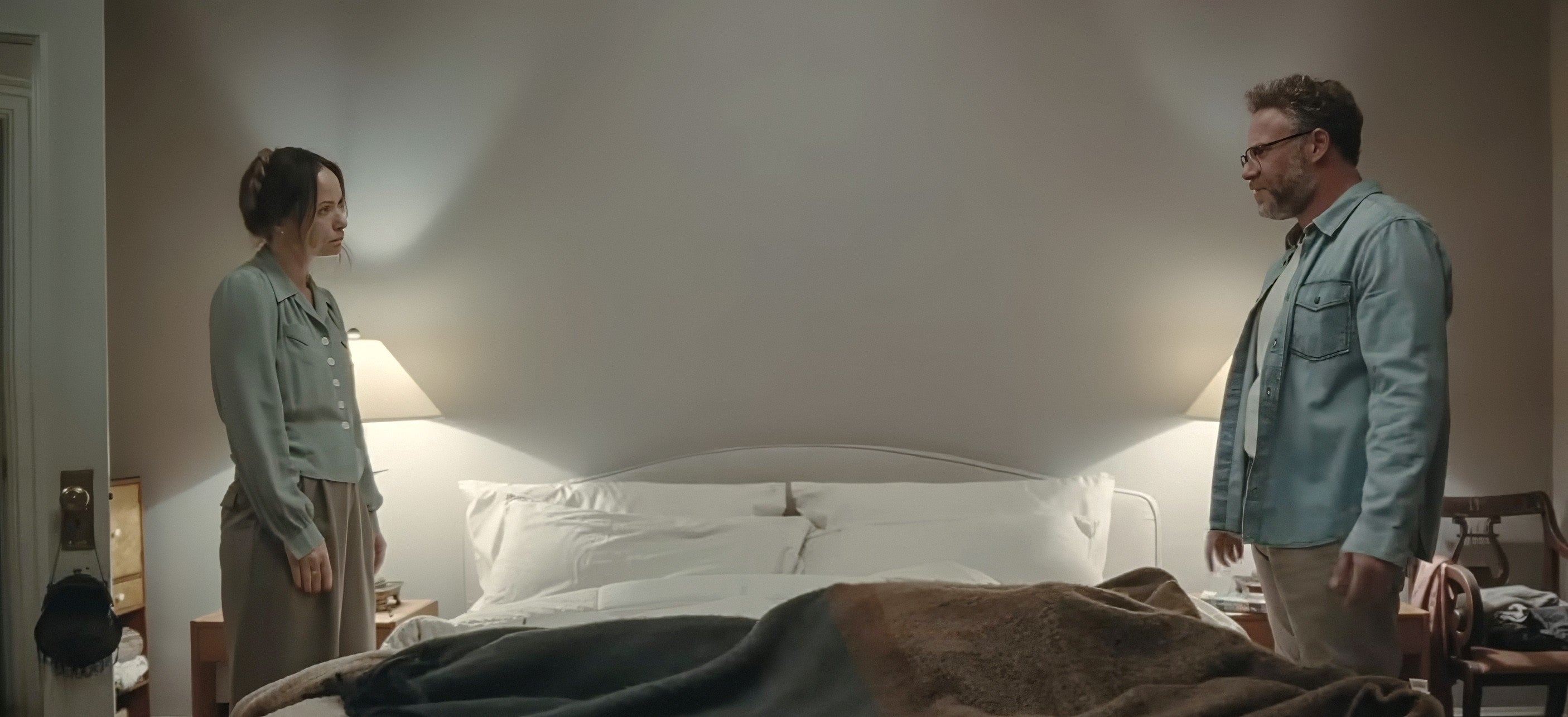 A man and woman stand on opposite sides of a bed in a dimly lit room, engaged in a tense conversation
