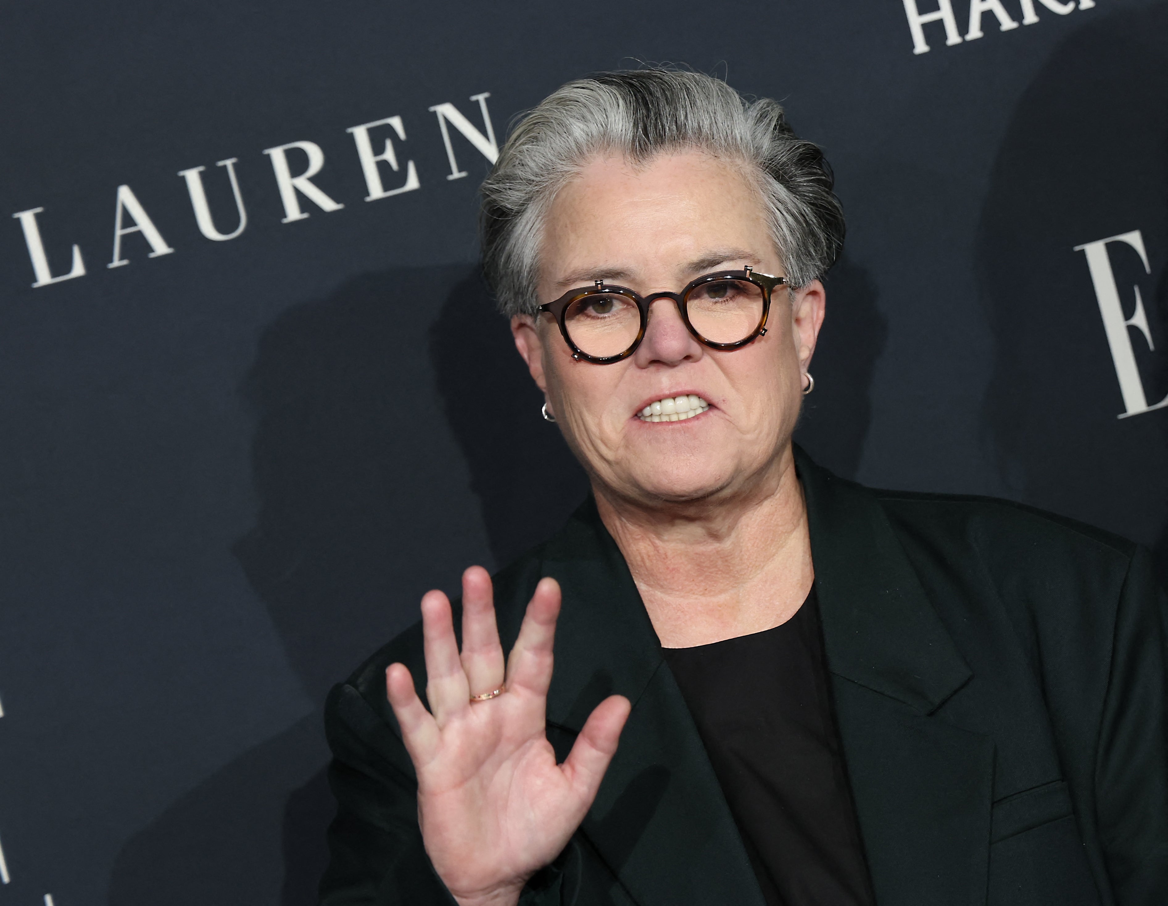 Rosie O'Donnell with short hair and glasses waves on the red carpet, wearing a blazer