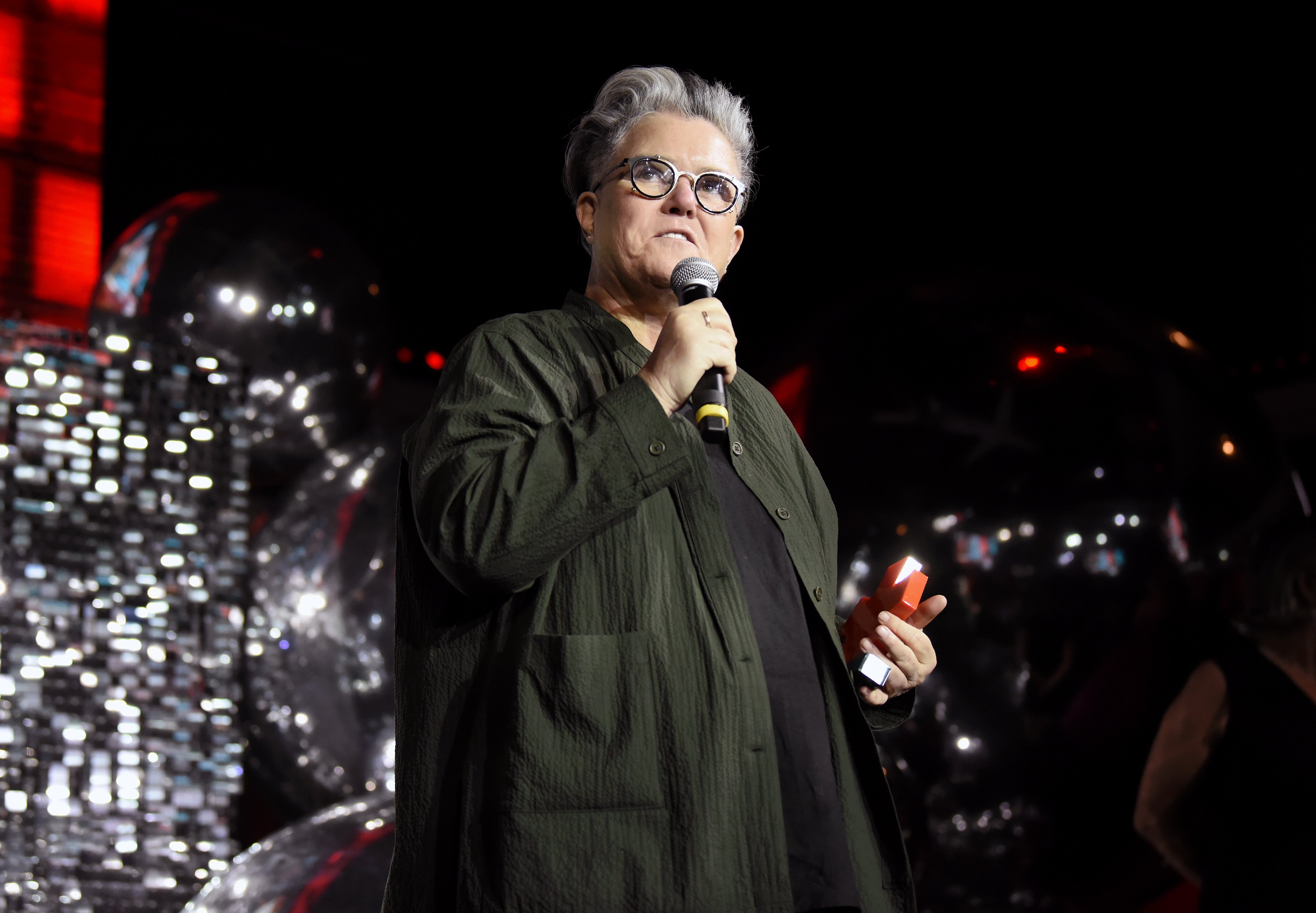 Rosie O'Donnell in a textured outfit speaks into a microphone on stage, holding a small item in the other hand, with a backdrop of shiny balloons