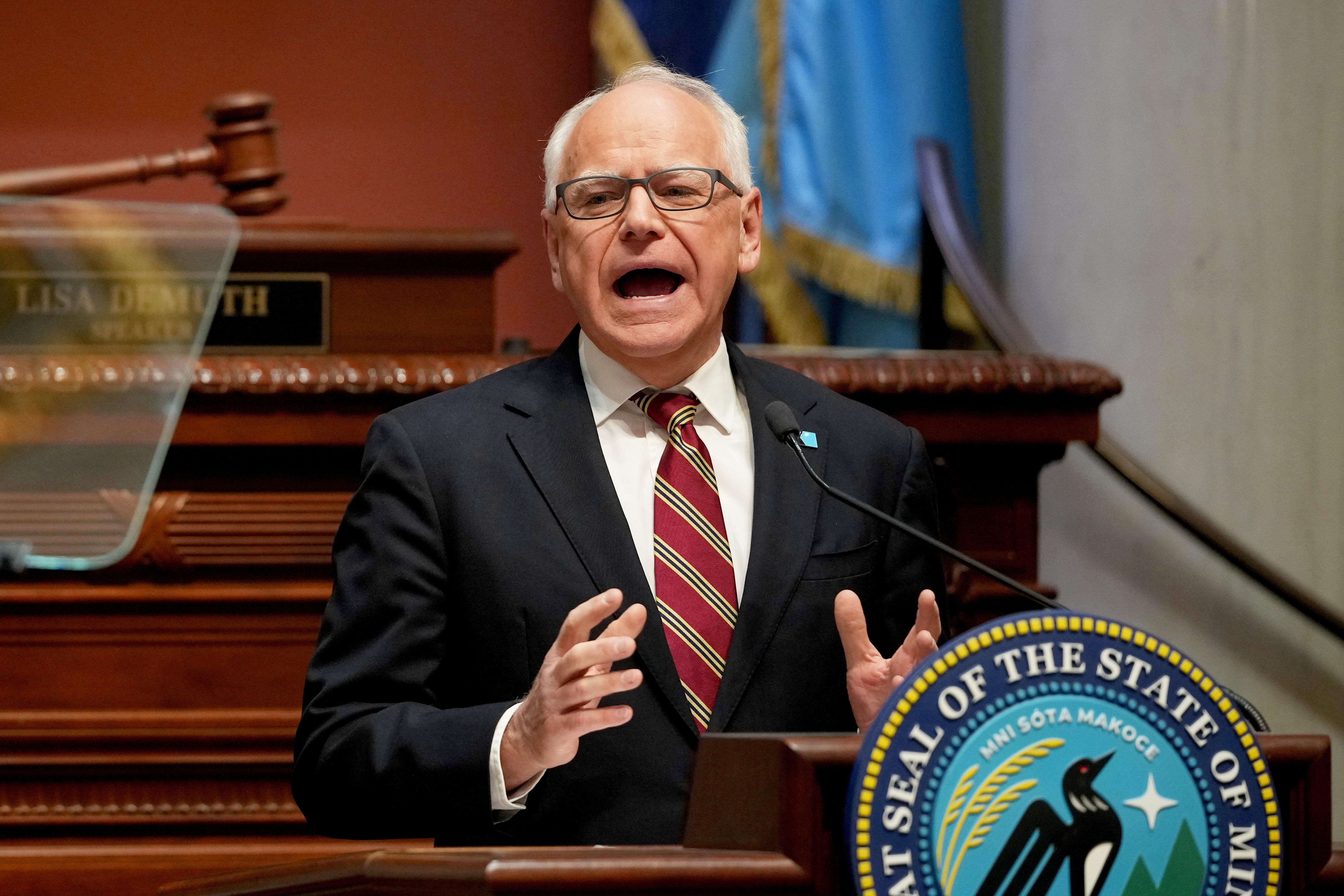 A man in a suit and striped tie speaks passionately at a podium with the State of Minnesota seal, inside a legislative chamber