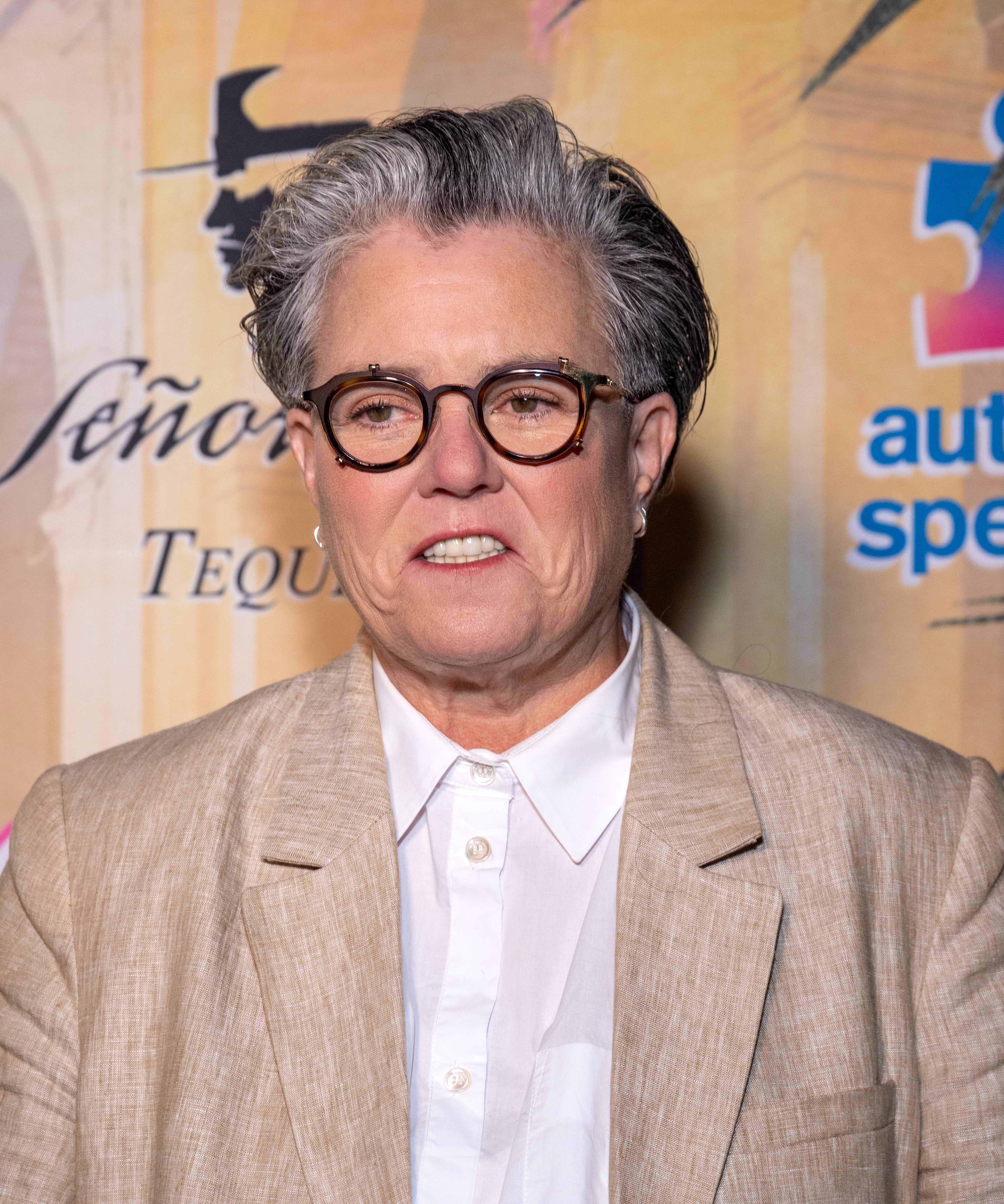 Rosie O'Donnell wearing a suit and glasses at a public event with logos in the background