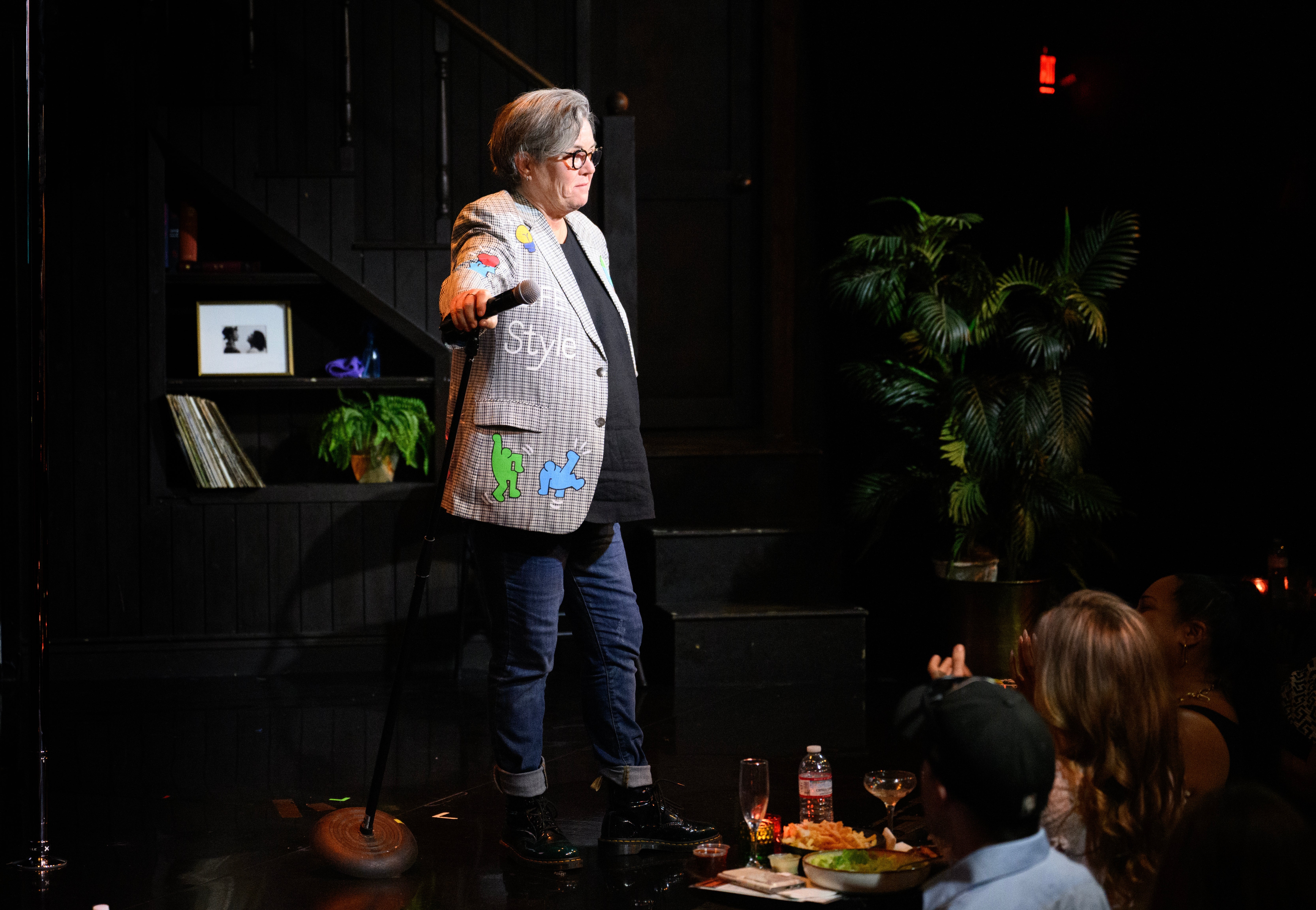 Rosie O'Donnell on stage, wearing a patterned blazer and jeans, holds a microphone while performing to an audience in a club setting