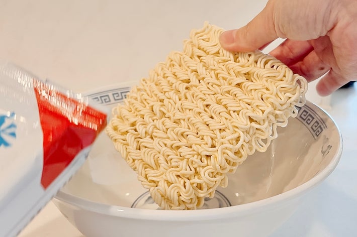 手がラーメンの乾麺をボウルに入れる瞬間。左には開封した袋が見える。