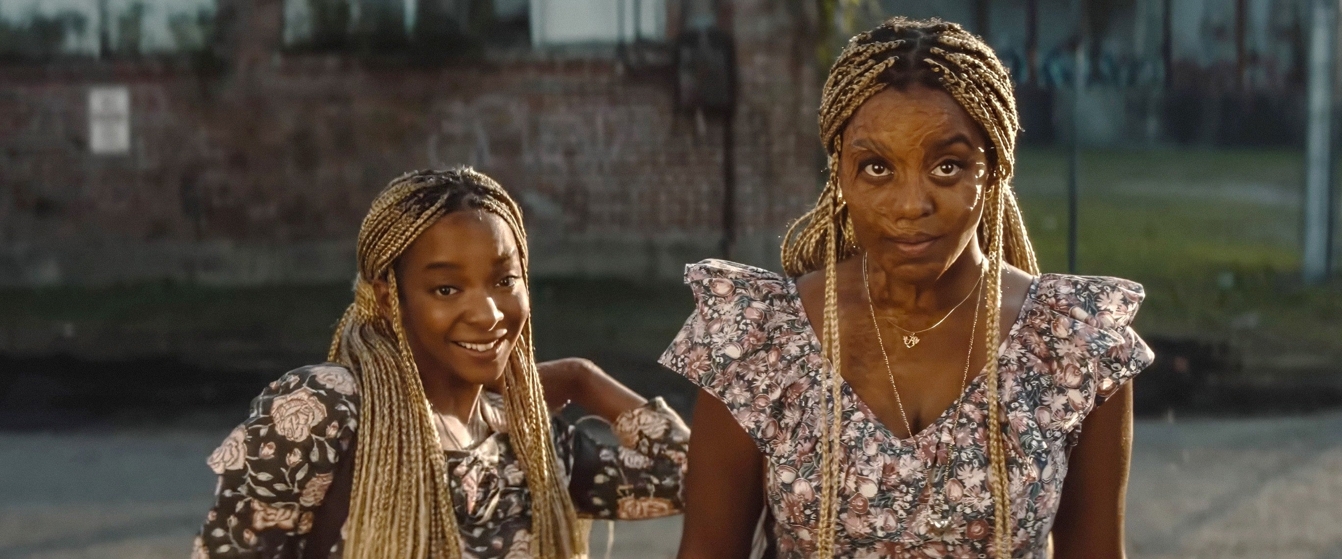 Two people with braided hair in floral tops. They stand outdoors, facing the camera with a relaxed expression