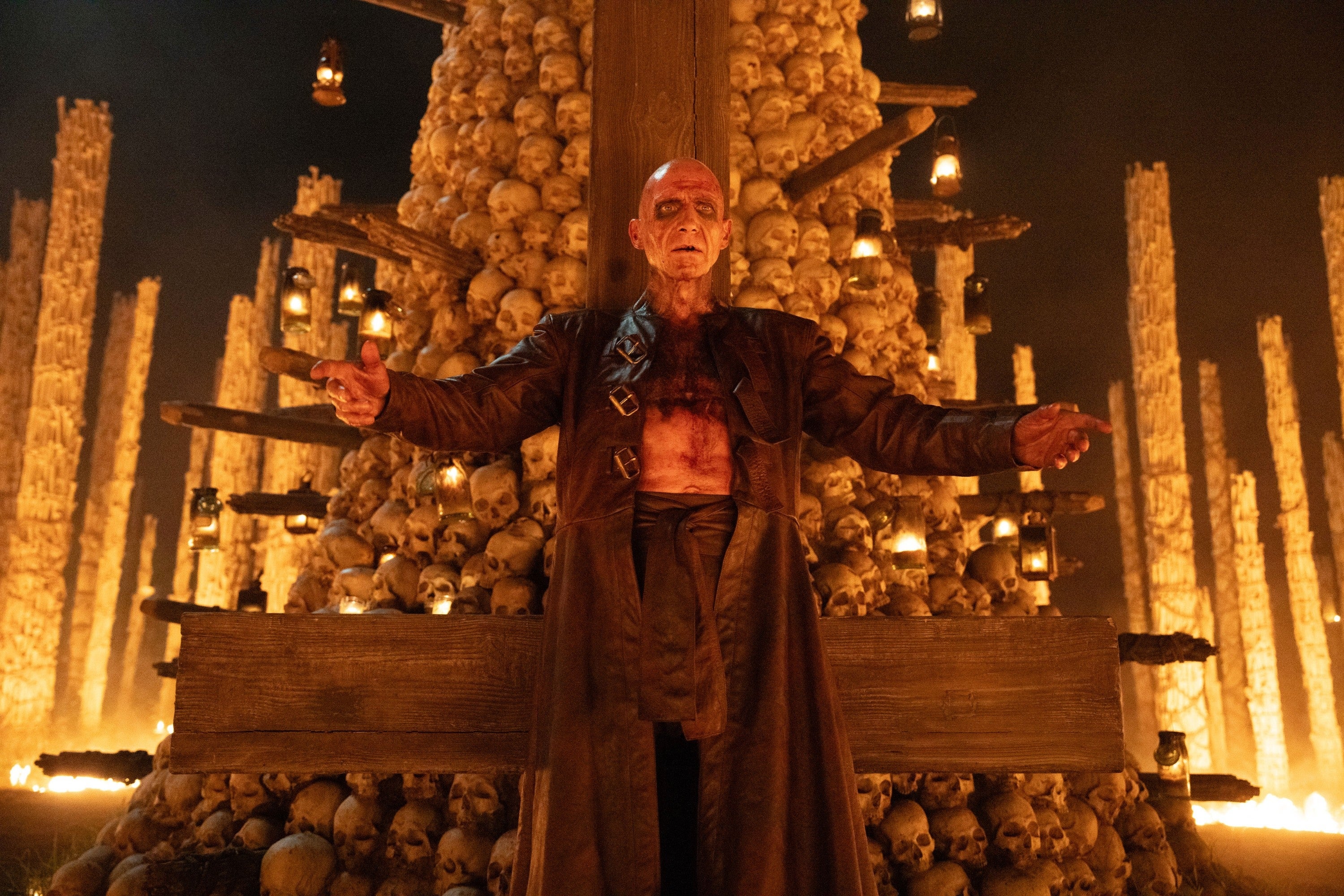 A character stands dramatically in front of a large pile of skulls with lit candles, arms outstretched, in a cinematic scene