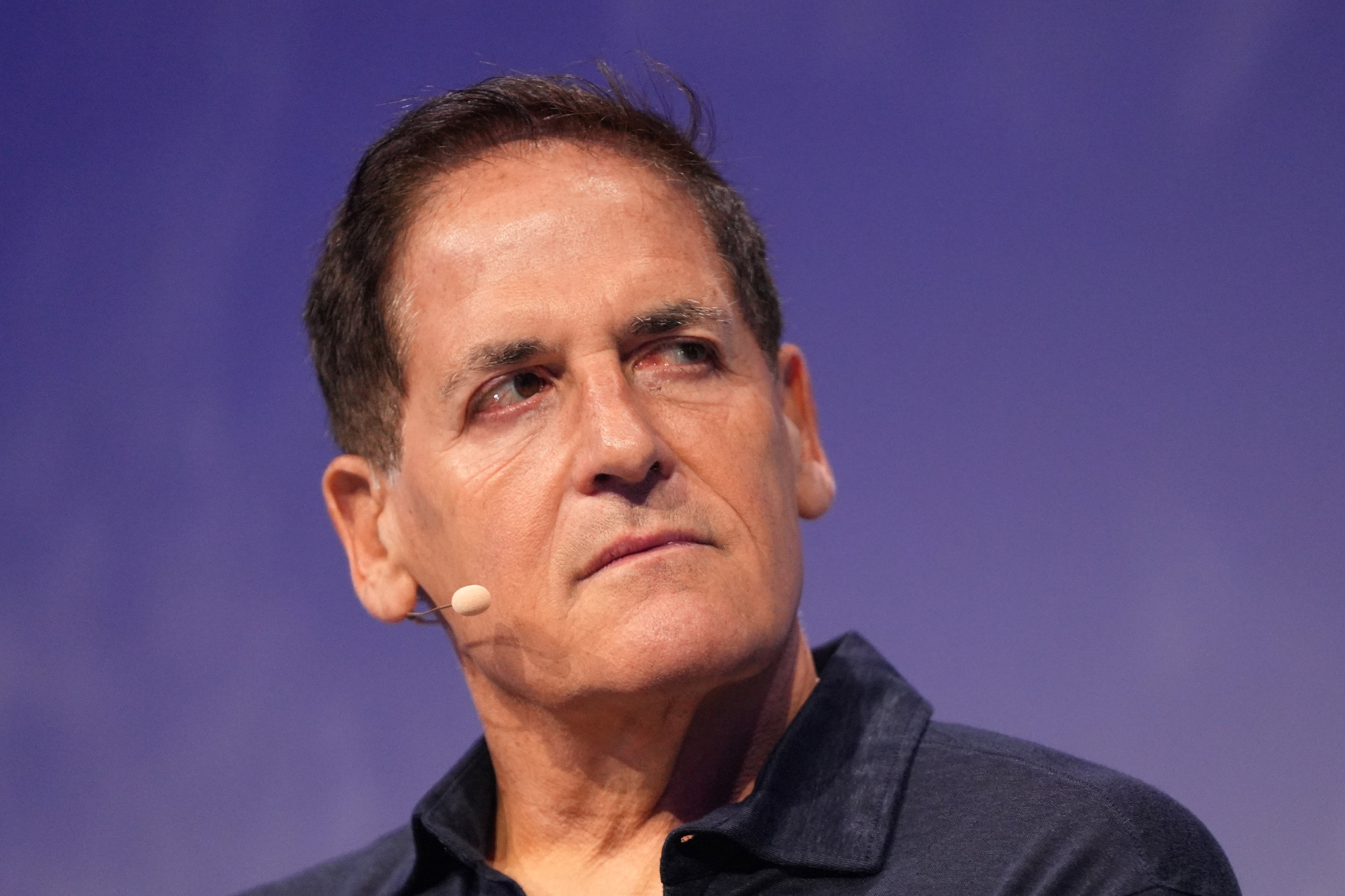 Mark Cuban with a microphone headset appears thoughtful against a plain backdrop, dressed in a casual dark shirt