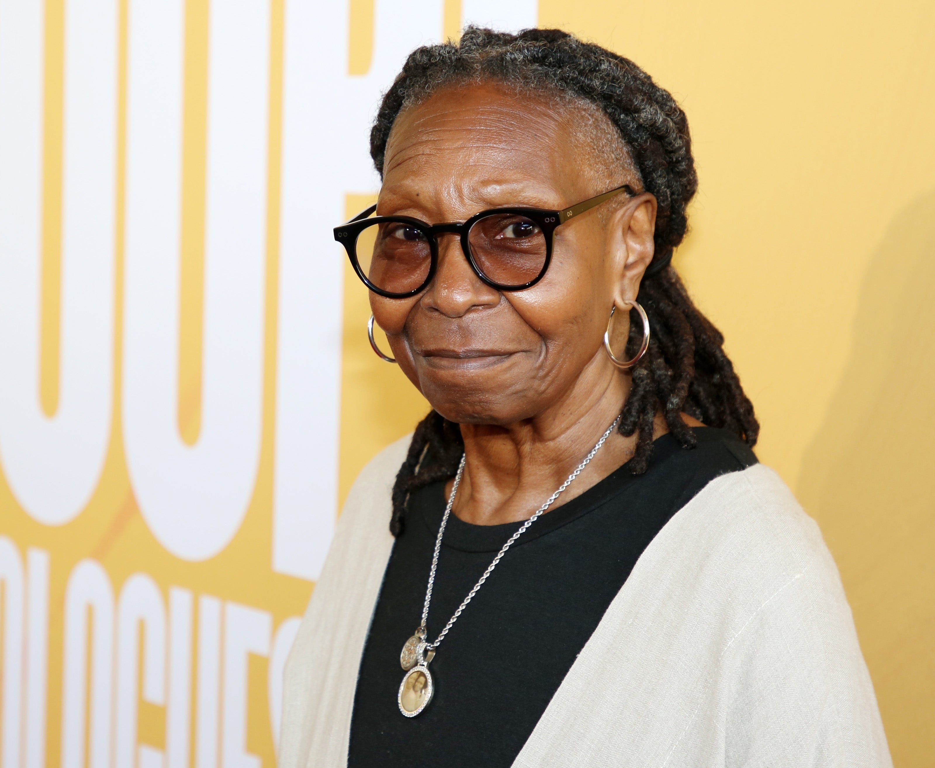 Whoopi Goldberg with glasses, wearing hoop earrings and layered necklaces, in front of a yellow backdrop with partial text visible