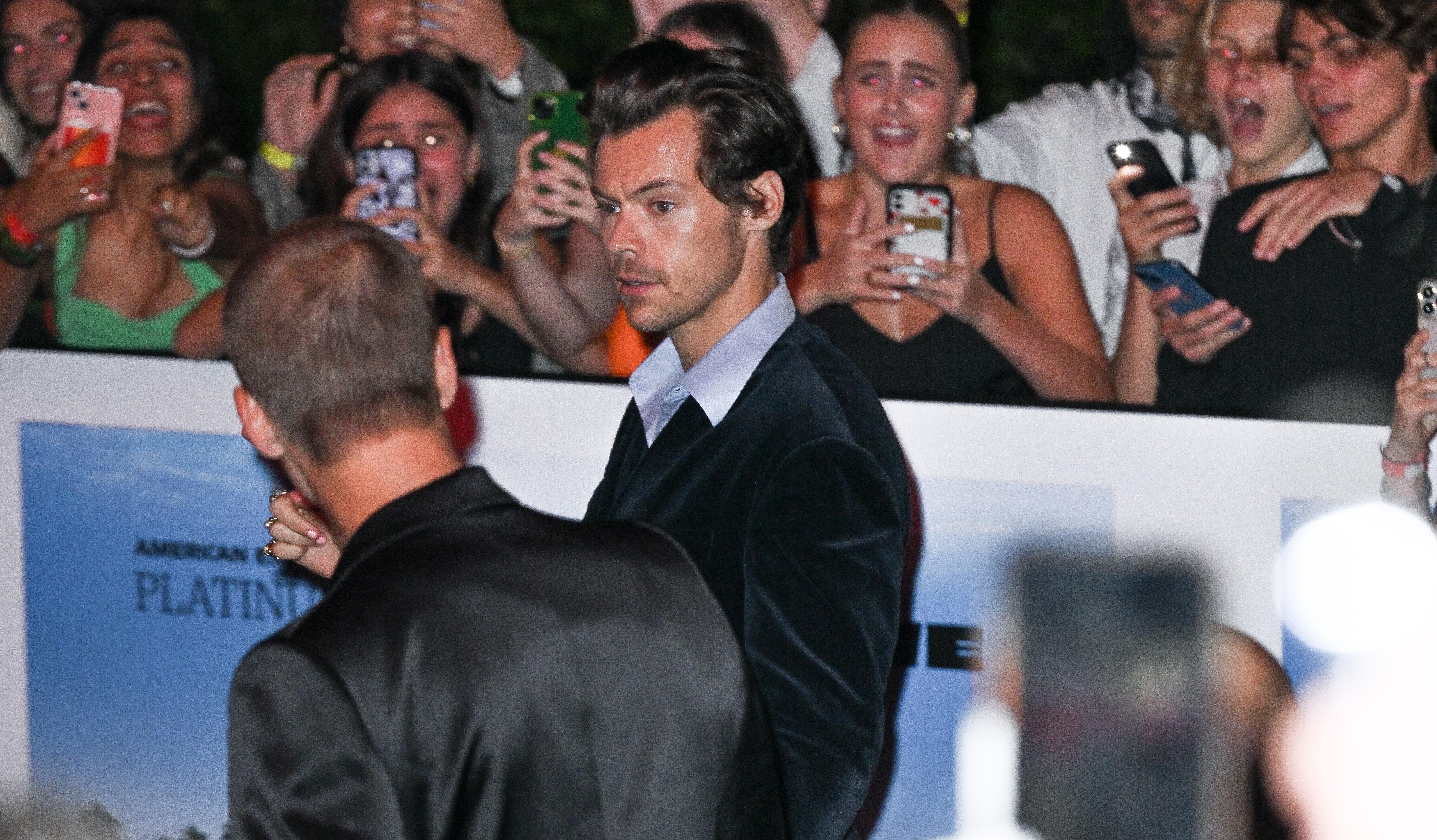 Harry Styles in a velvet suit at an event, with fans excitedly taking photos in the background