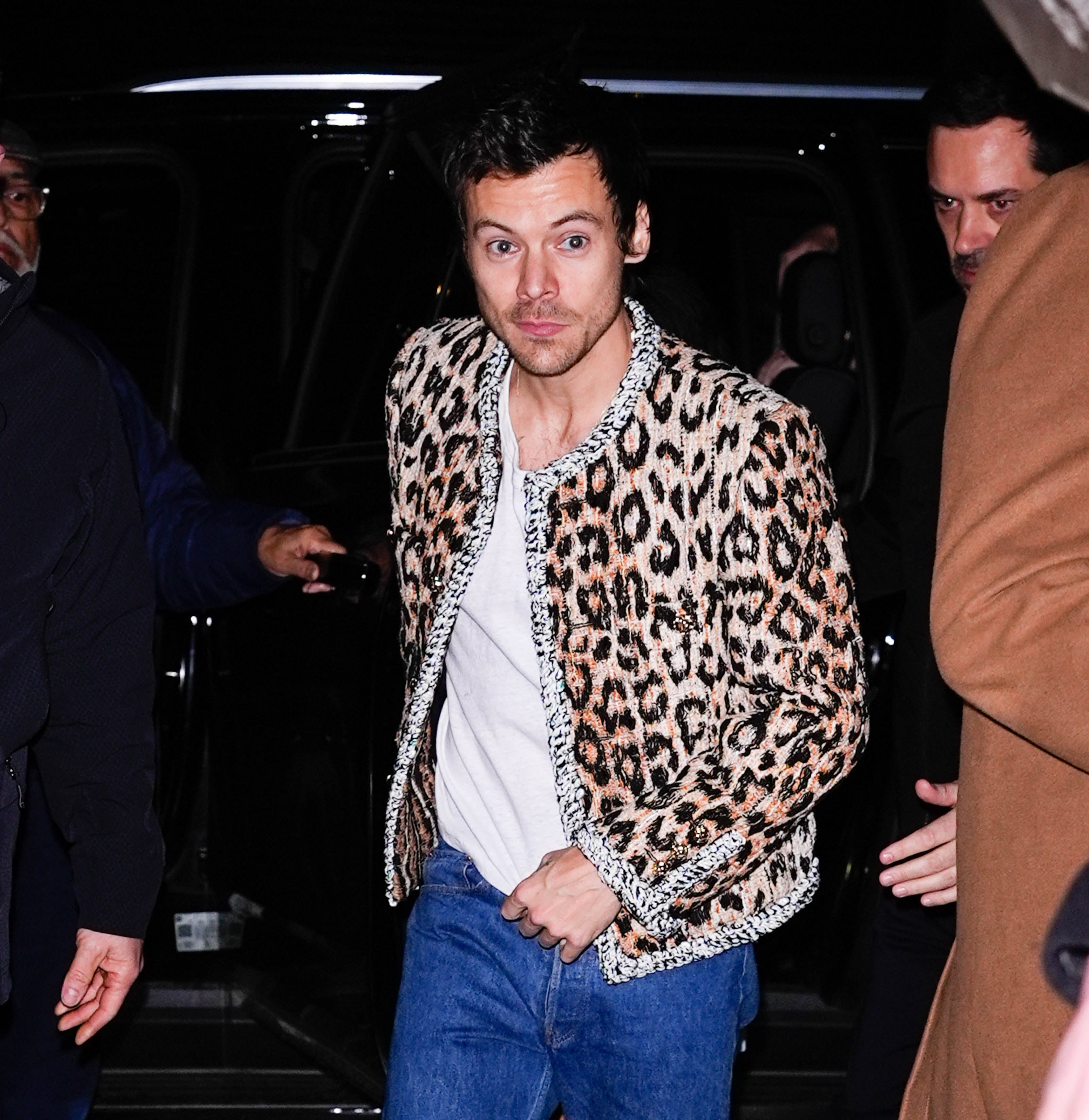 Harry Styles in a leopard print jacket, shirt, and jeans, walking outside with a group in the background