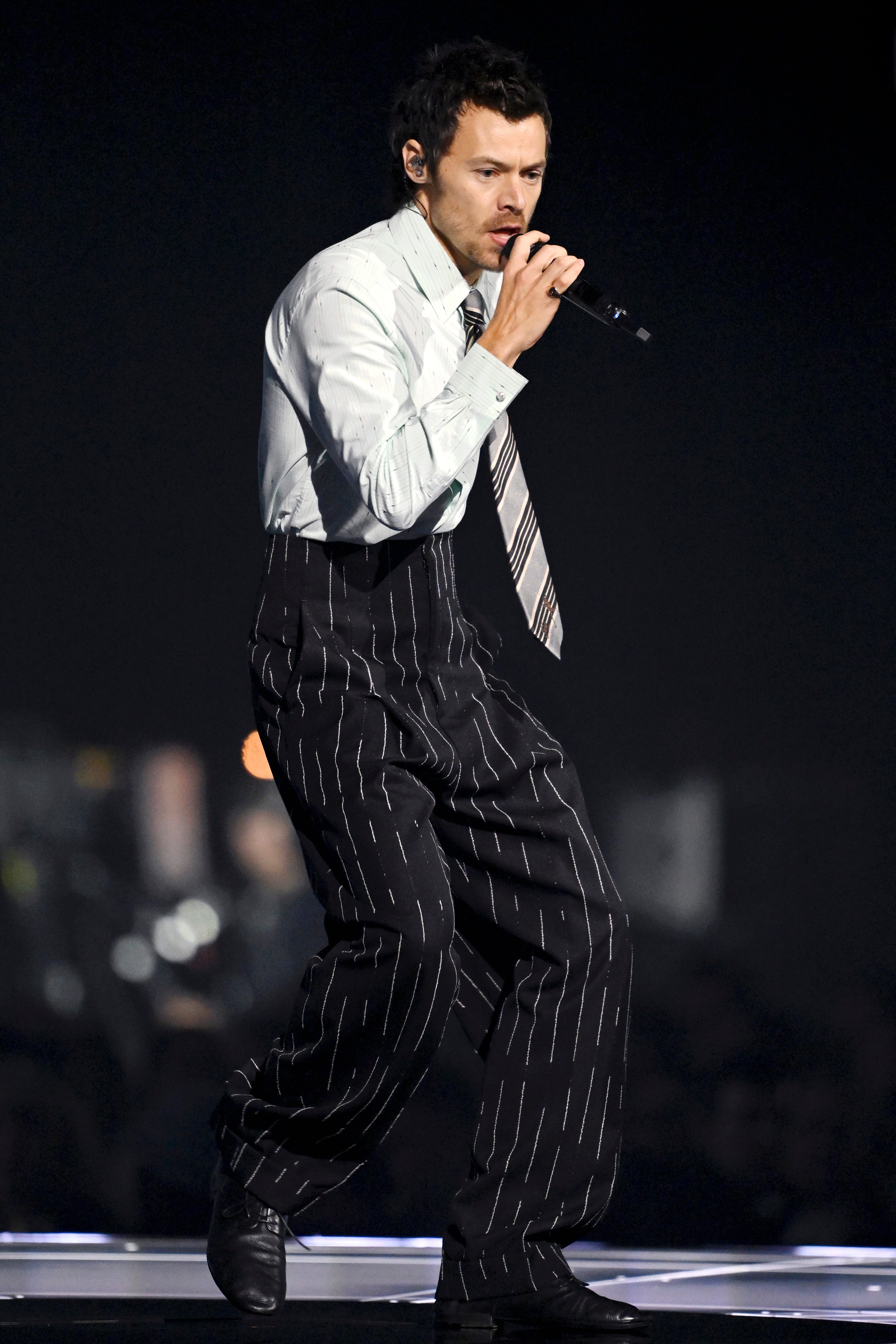 Harry Styles in pinstripe suit and tie performing on stage with a microphone