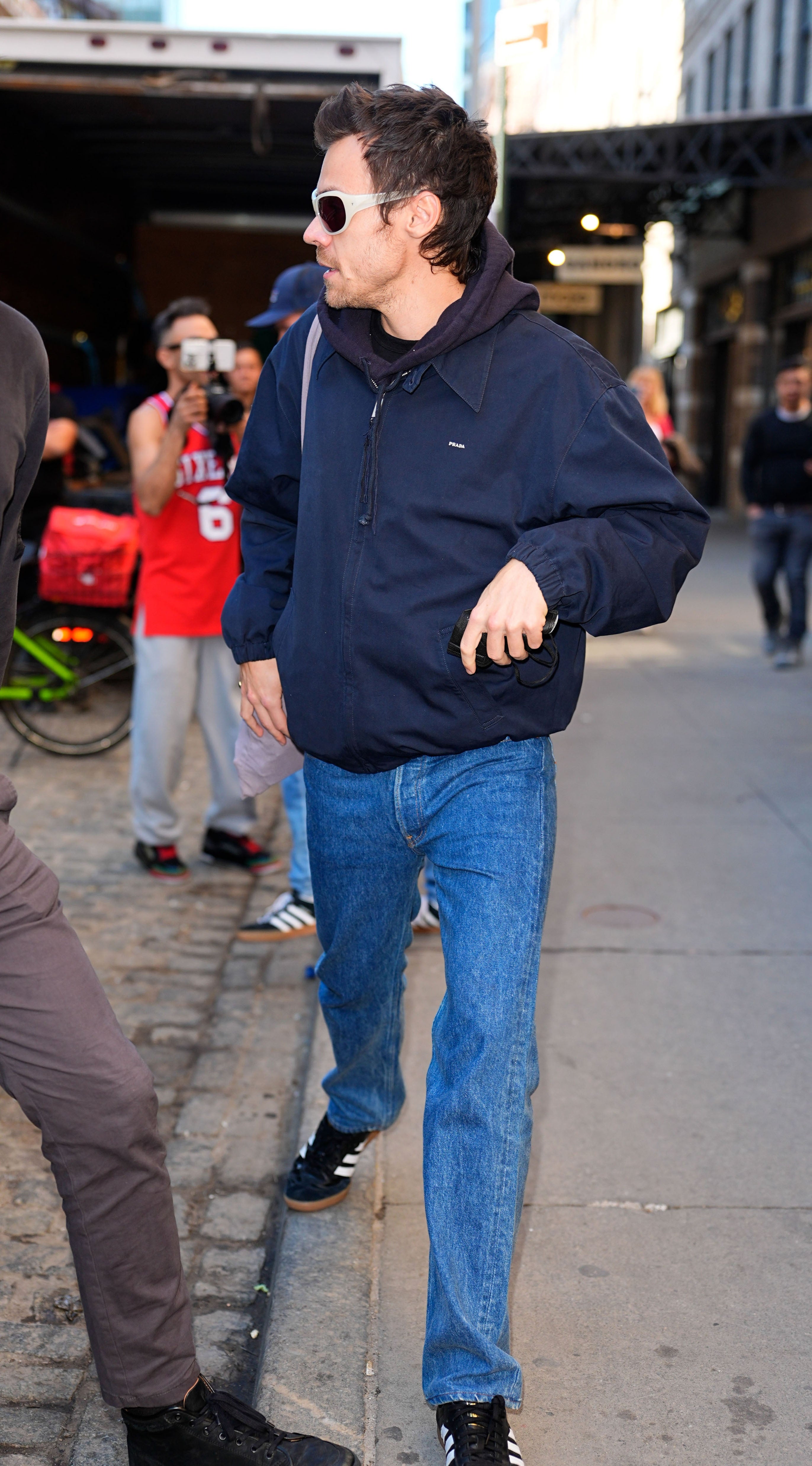 Harry Styles in casual jeans, a jacket, and sunglasses walks on a busy street, surrounded by onlookers