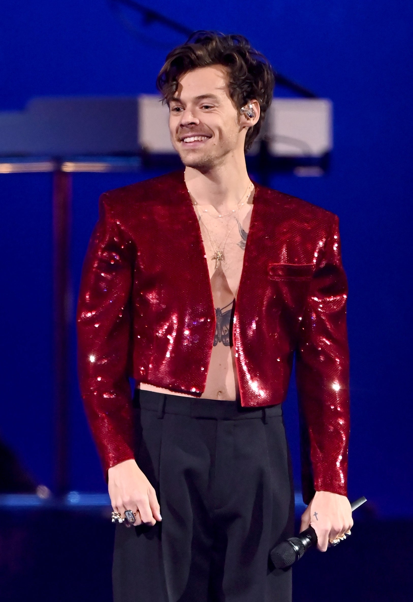 Harry Styles in a sequined jacket and pants holds a microphone on stage, smiling, with instruments in the background