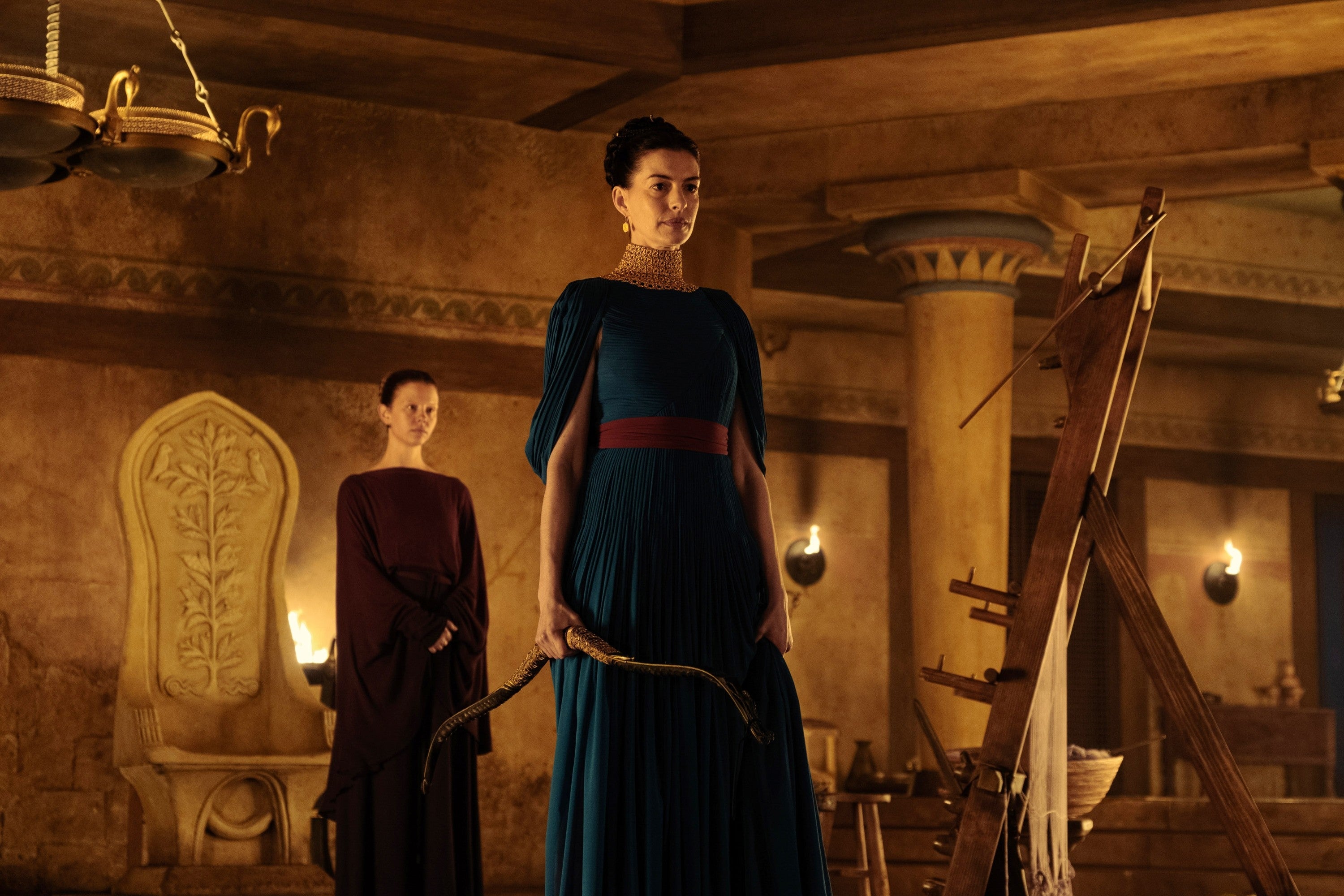 Two women in an ancient, dimly-lit room. One holds a bow, dressed in a long, elegant gown, while the other stands behind, observing quietly