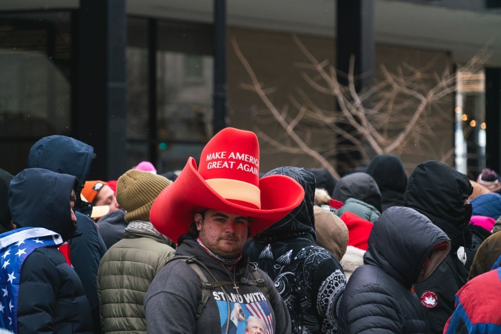 Person i stor rød cowboyhatt med "Make America Great Again" tekst står i en folkemengde. De har på seg jakke og hettegenser, vendt fremover