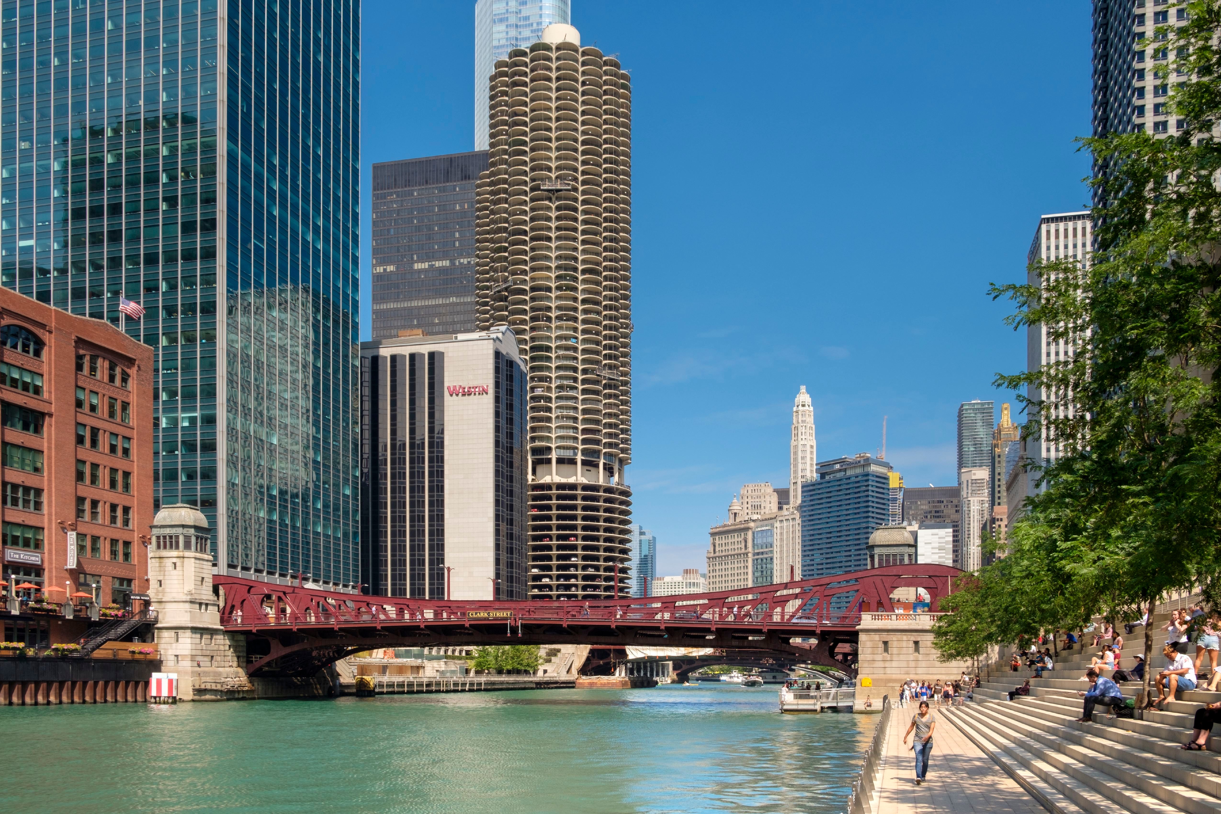 Utsikt over en elvebredd i Chicago med skyskrapere, en rød bro og folk som sitter langs trappen ved elvebredden