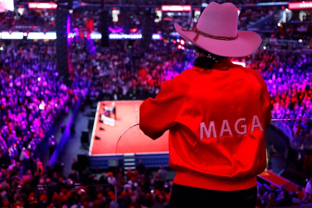 Person i cowboyhatt og "MAGA" jakke har utsikt over et overfylt stadionarrangement med en høyttaler på scenen