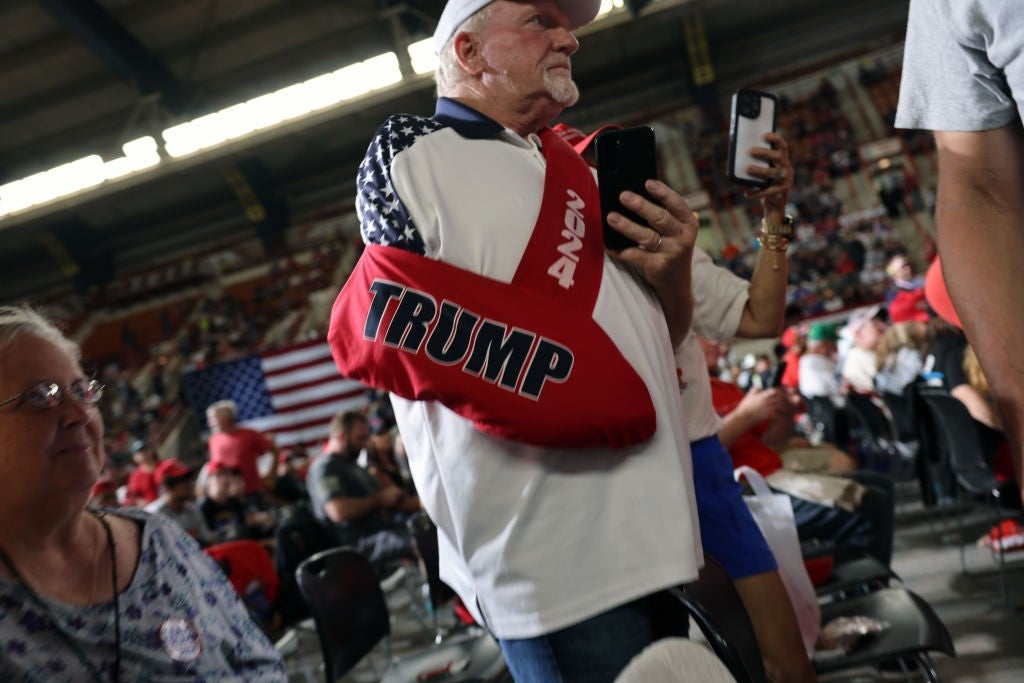 Mann med "Trump" slynge og hatt på et overfylt innendørsarrangement, med en telefon