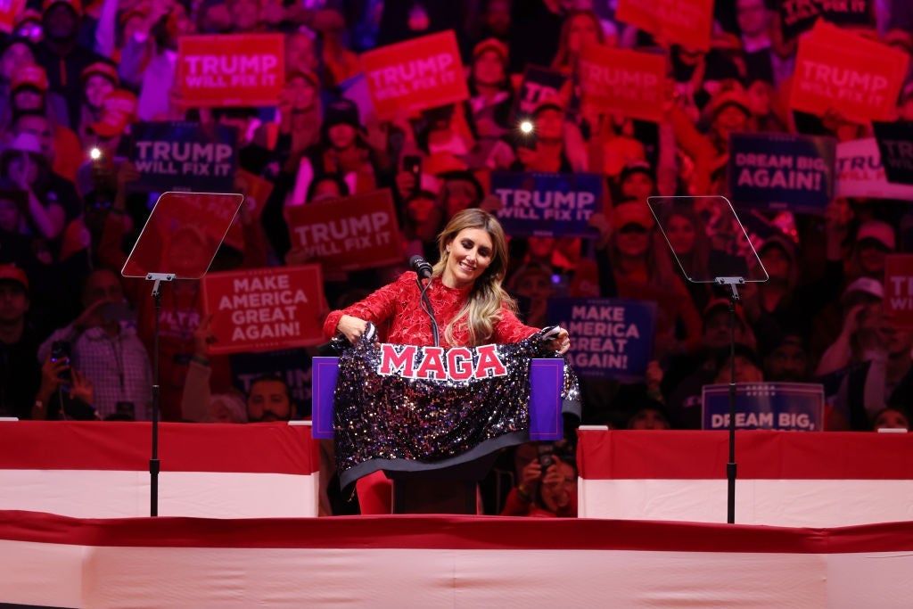 En kvinne i en paljettjakke holder en "MAGA" banner på et møte, omgitt av supportere med skilt
