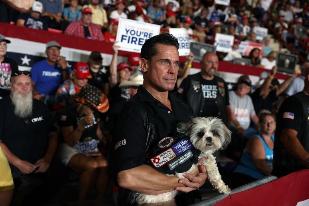 Mann i en overfylt begivenhet som holder en liten hund, omgitt av folk som holder "You're Fired" tegn