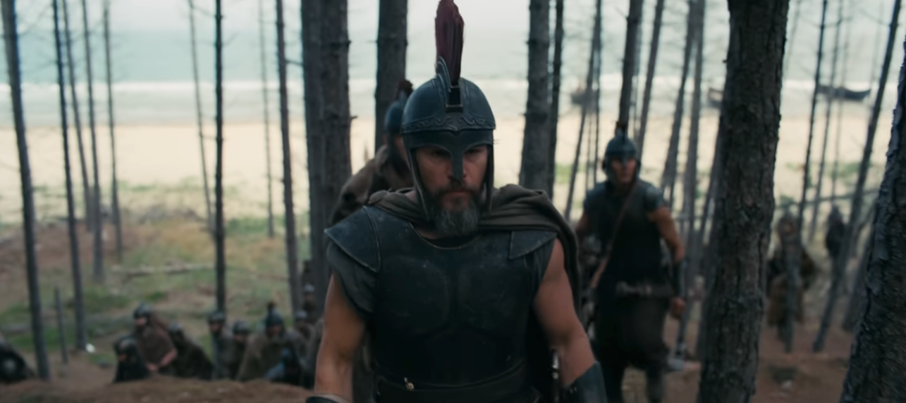 A group of armored warriors with helmets navigate through a forest near a beach, led by a determined figure at the front