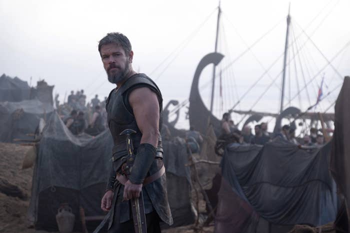 A warrior in armor stands on a beach with Viking ships in the background, appearing serious and ready for battle