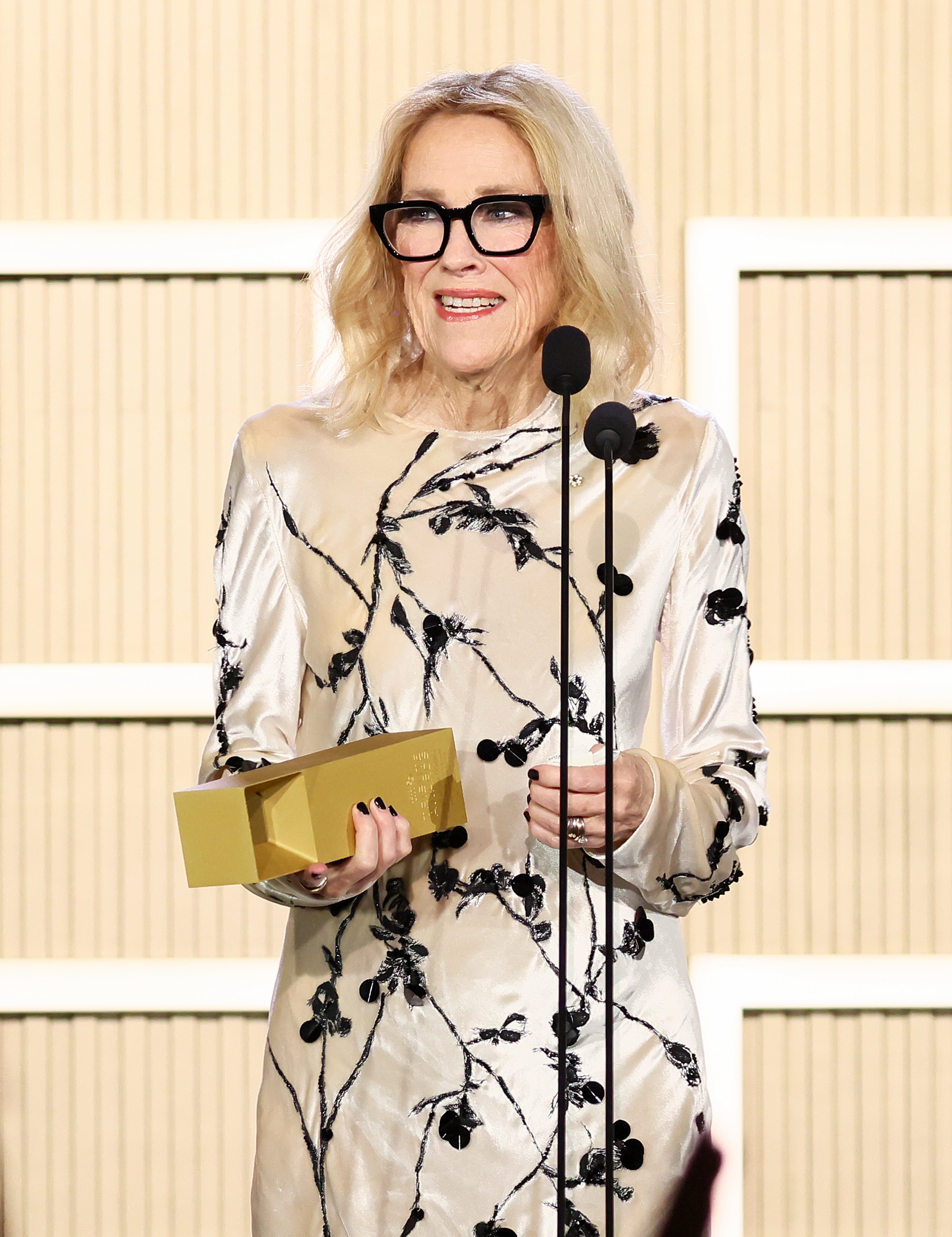 A person in a stylish embroidered gown and glasses stands on stage holding a gold award while speaking at a microphone
