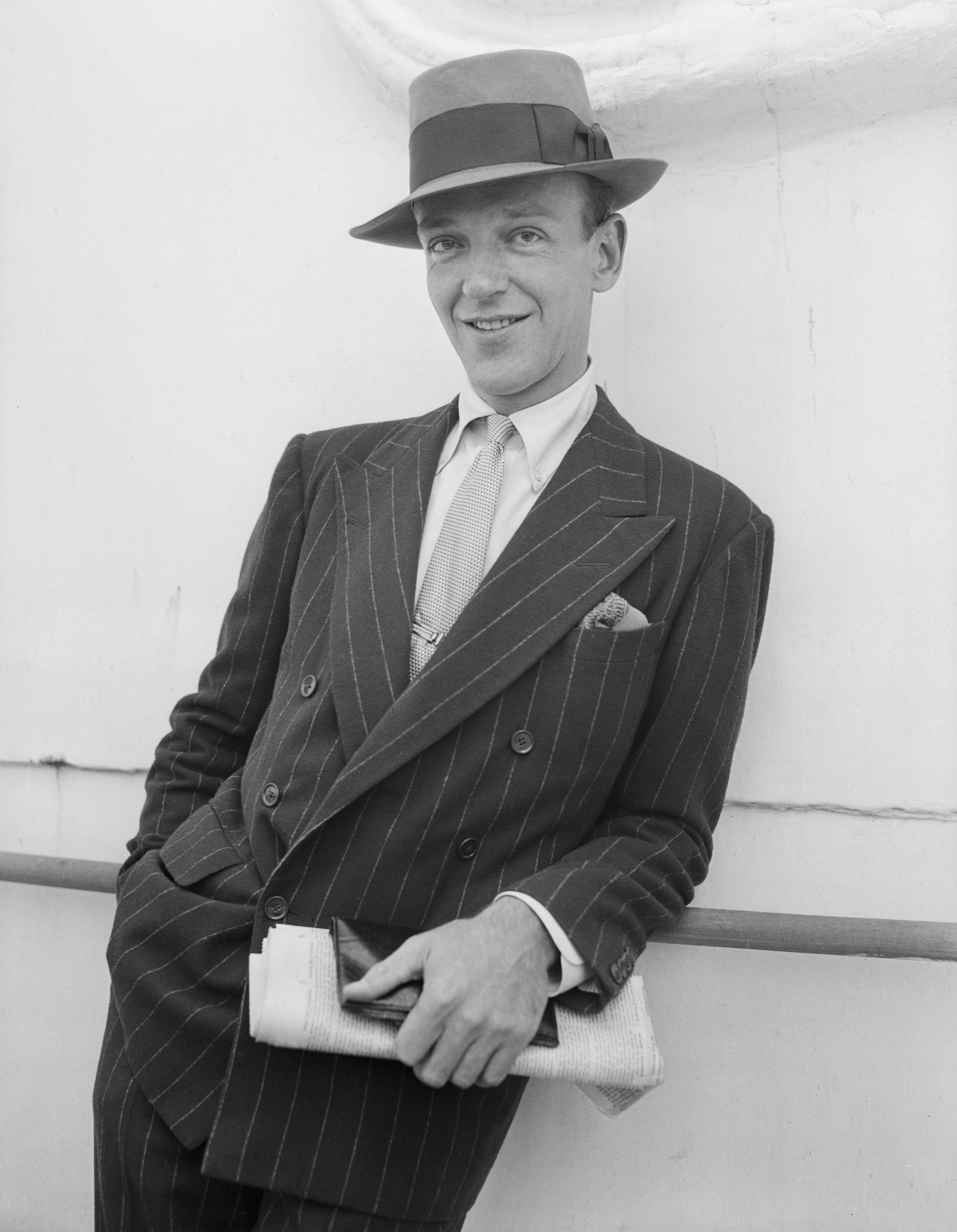 Fred Astaire in a pinstripe suit and fedora, holding a rolled paper and leaning casually against a railing, setting a vintage, classic style