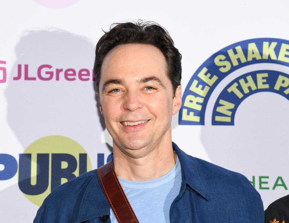Person smiling at an event, wearing a casual blue jacket over a light blue shirt. Background shows partial text and logos