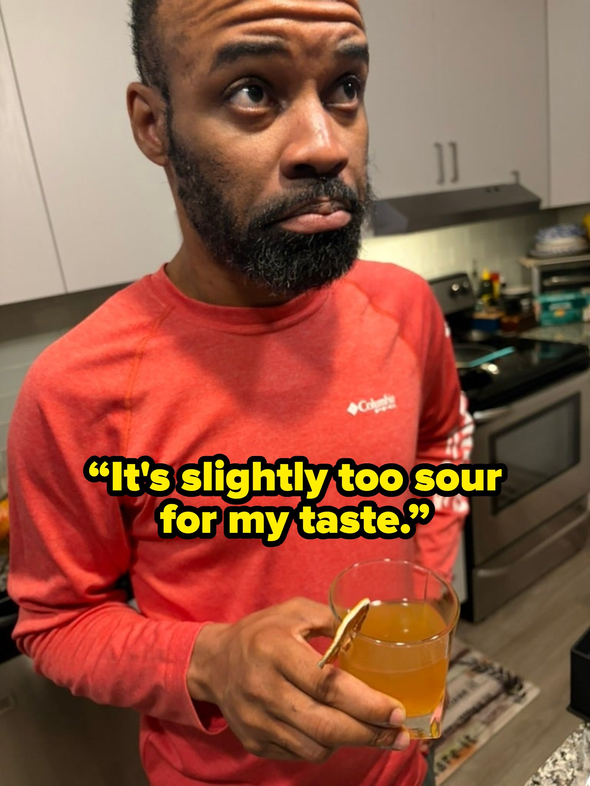 Person in a kitchen holds a glass of tea with a slice of lemon, looking contemplative