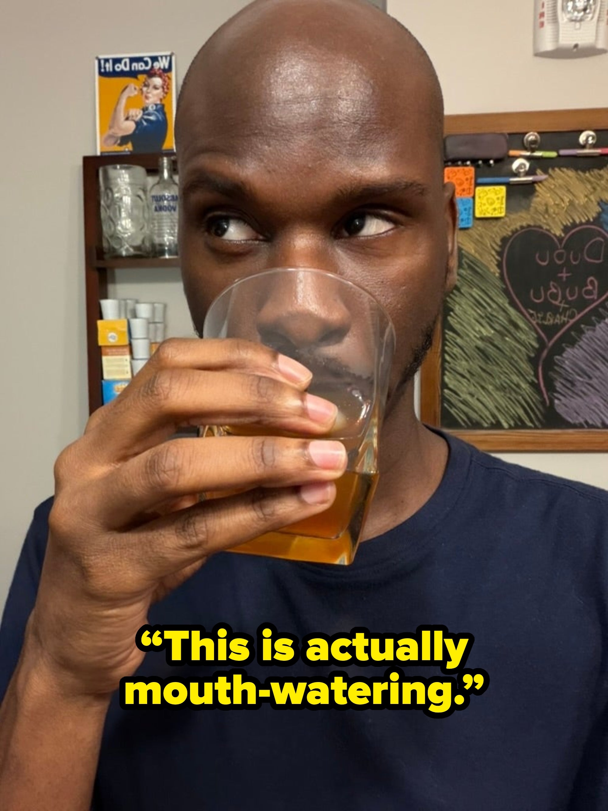 Person drinking from a glass, looking sideways in a kitchen. Bulletin board with notes is visible in the background