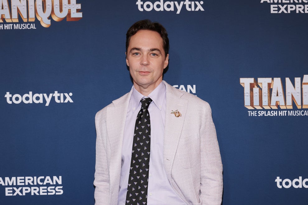 Person in a light suit and patterned tie attends a theater event, standing in front of a backdrop with event branding
