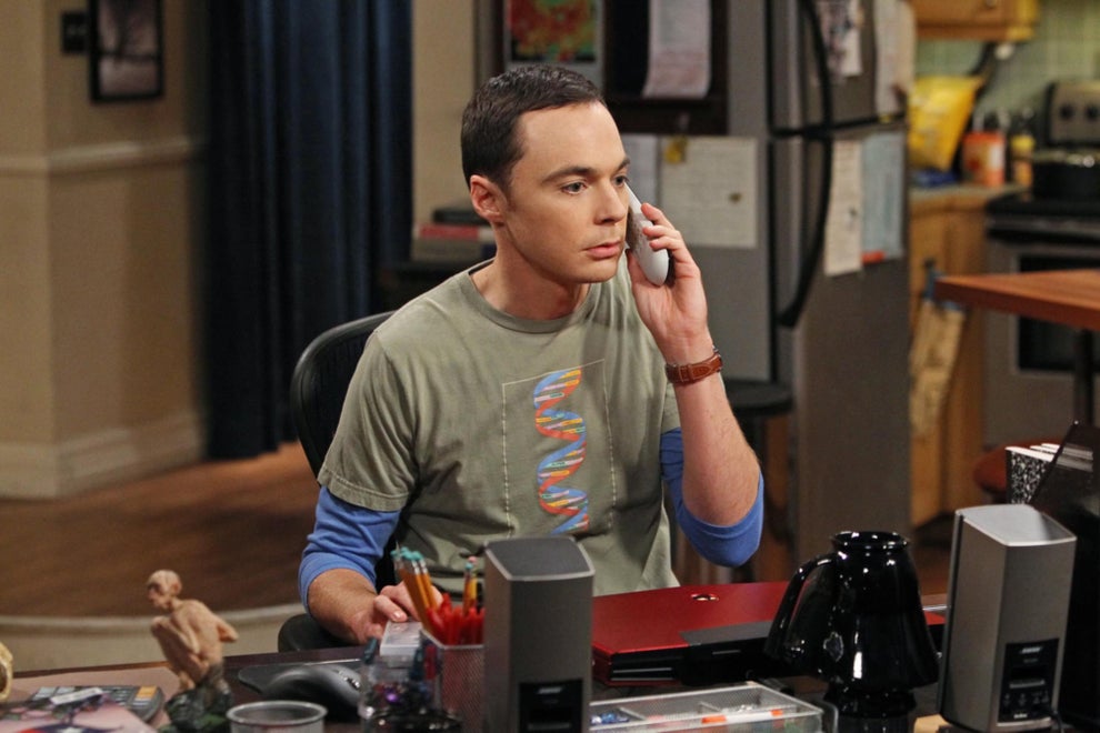 Person sitting at a cluttered desk, holding a phone to their ear, appearing focused. Casual layered clothing; household setting in the background