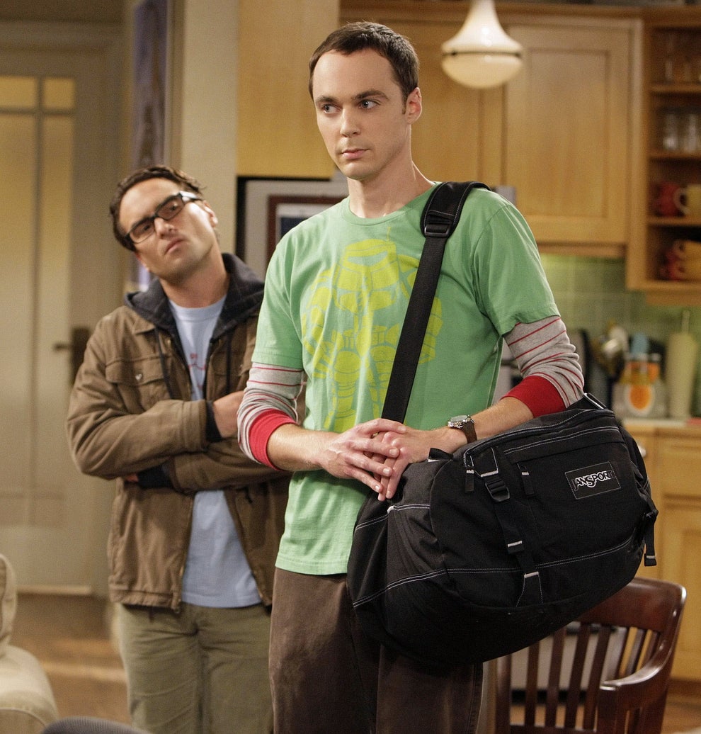 Two people in a living room; one wears a green T-shirt with a bag, the other wears glasses and a jacket with folded arms